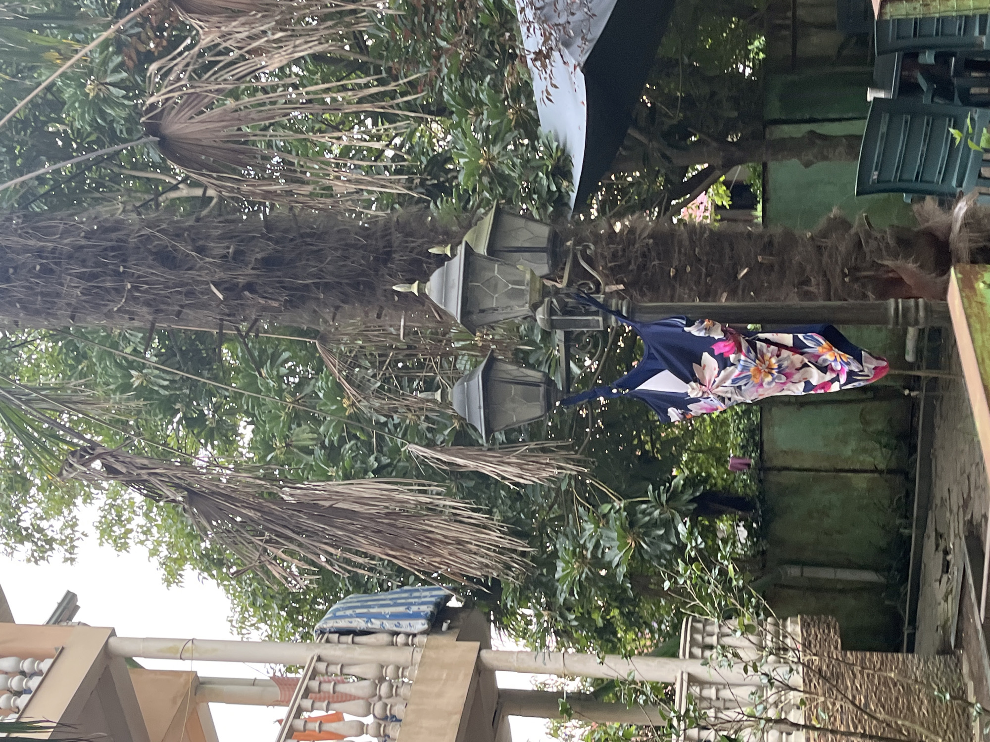 In Ureki, Georgia, a swimsuit with a floral pattern hangs from a lamppost in a courtyard. The lamppost is situated next to a palm tree with dry fronds. In the background, a building with a balcony is visible, with laundry items, including a striped blue and white blanket, draped over the railing. The courtyard contains green plastic chairs and a dark umbrella. The scene appears to be outdoors, with lush green foliage surrounding the area.