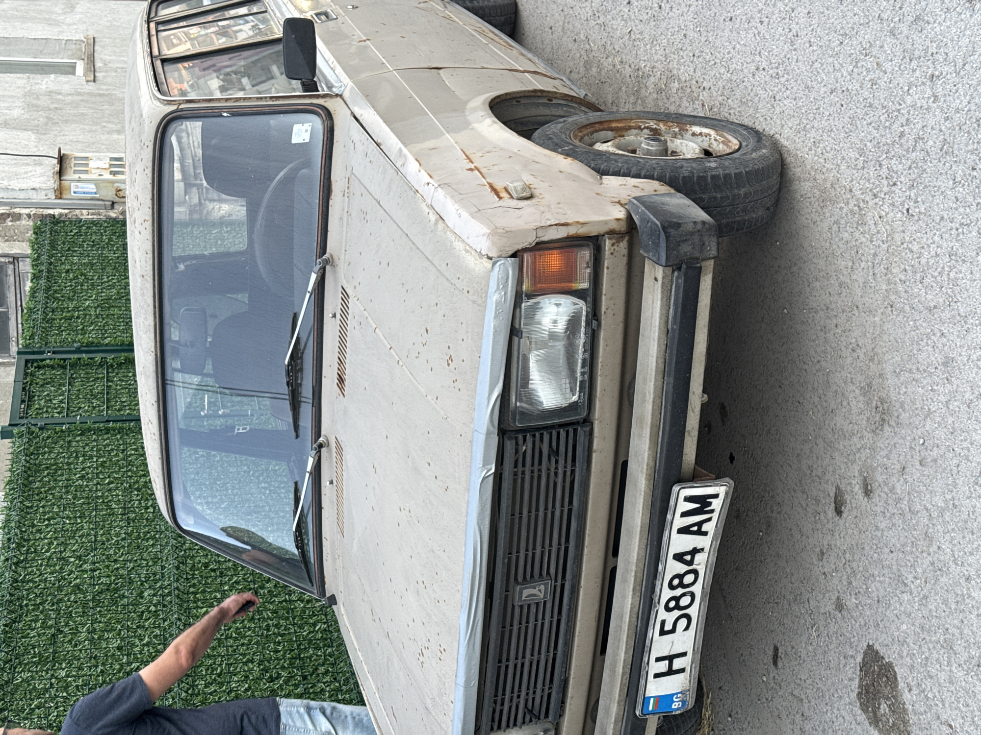 This image, rotated 90 degrees counter-clockwise, depicts a heavily rusted and weathered beige Lada Classic series car (likely a 2105 or 2107 model) parked on an asphalt road in an urban environment. The vehicle exhibits significant signs of age and neglect, with extensive rust covering its body, particularly on the hood and fenders, and peeling paint. A spare, equally worn tire with a rusty rim is visible resting on the front left fender area, partially obscuring the wheel well.
In the lower left foreground, a person's arm and torso are visible, clad in a dark t-shirt and light blue jeans, with their hand appearing to reach towards the car's windshield or bonnet. The car's front features its distinctive black plastic grille with the Lada logo, a single amber turn signal, and a clear headlight visible on the right side. Black plastic bumpers are also present.
The front license plate clearly displays "H 5884 AM" with a small "BG" country code and the European Union flag, indicating it is from Bulgaria, specifically the Shumen region (identified by the 'H' code).
The background includes a green artificial foliage fence directly behind the car, and a light-colored concrete or stucco building further back on the left. The scene is illuminated by daylight, suggesting a clear or overcast day. The car appears to be in a state of disrepair, potentially abandoned or infrequently used, against a backdrop of common urban elements in Shumen, Bulgaria.