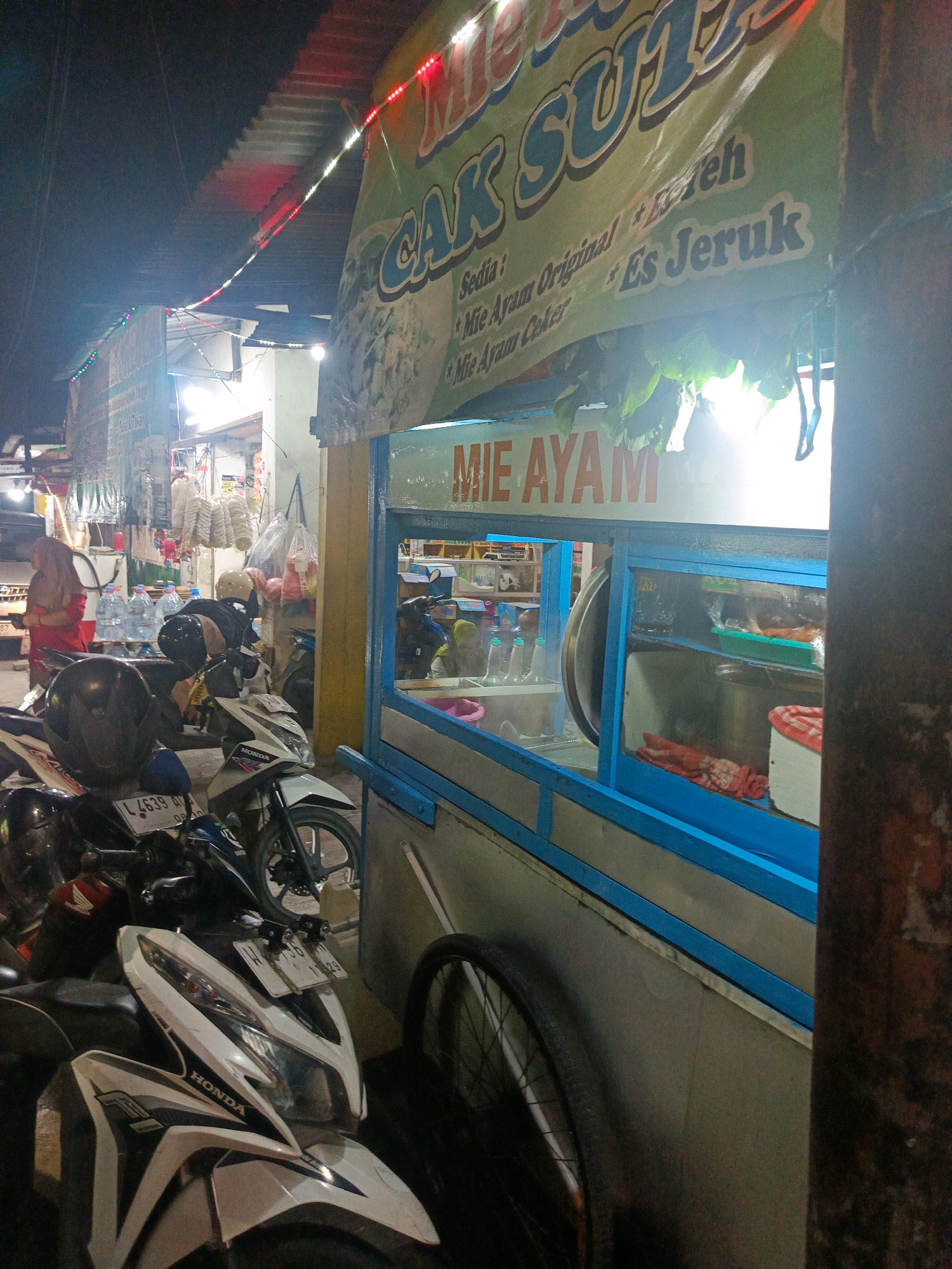 This image shows a nighttime scene at a food stall in Sedati Agung, Indonesia. A banner with text is visible, indicating "MIE AYAM" (chicken noodles) and "Es Jeruk" (orange ice). The stall appears to be a mobile cart with a blue frame and a glass display. Several motorcycles are parked in front of the stall, some with helmets on them. In the background, there are more stalls and what appear to be goods displayed for sale. The lighting suggests it is dark, with artificial lights from the stall and string lights illuminating the area. A person in a red hijab is visible in the background near the motorcycles. Another person, possibly the vendor, is leaning over a motorcycle, wearing dark clothing and a helmet. The overall atmosphere is that of a busy street food market.