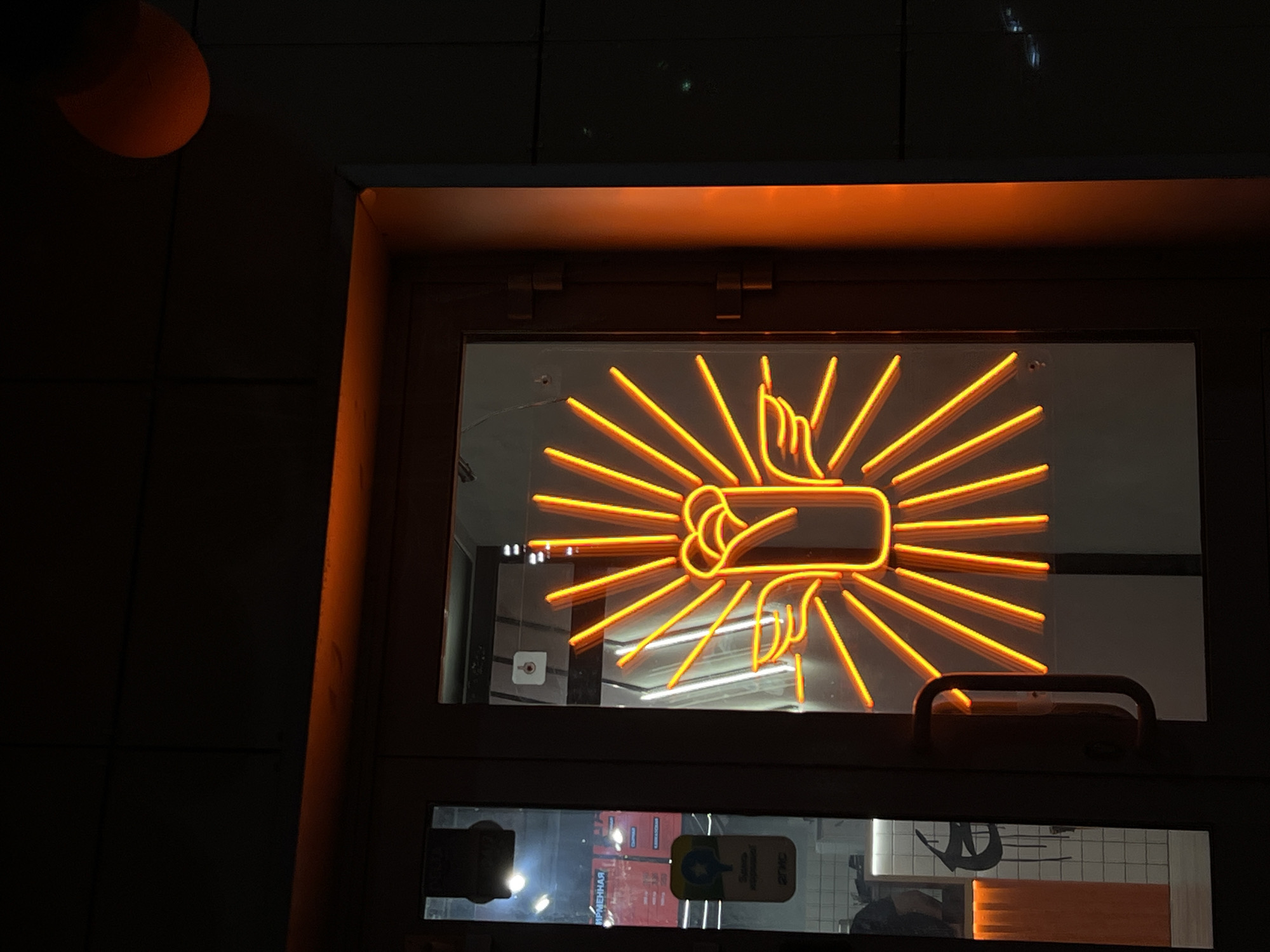 The image shows a neon sign in a window. The sign depicts a fist making the "fig sign," or "shish" gesture, with radiating lines around it. The sign is illuminated in a bright orange color. The window is set into a frame that is also lit with orange light. Through the window, glimpses of the interior of a building can be seen, including what appears to be a tiled wall and other signs. Based on analysis of internal information it is likely this image was taken in the city of Kazan, Russia.