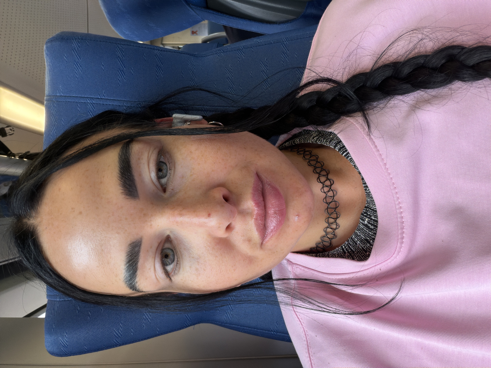 The media file contains a close-up, low-angle, portrait-oriented shot of a young woman lying down. She has dark hair styled in a braid that rests over her shoulder and chest. She is wearing a light pink crewneck sweatshirt over a silver, textured top, and a black choker necklace. Her face is turned towards the camera, with her left eye more prominent. She has a light dusting of freckles across her nose and cheeks. Her lips are full and appear to have a light pink or nude lipstick.
The background is blurred but suggests an indoor setting with blue upholstered seating, possibly on a train or bus. The lighting is bright, indicating daytime. The woman's expression is neutral. There is no visible text in the image. The setting appears to be Ушаки, Russia.
