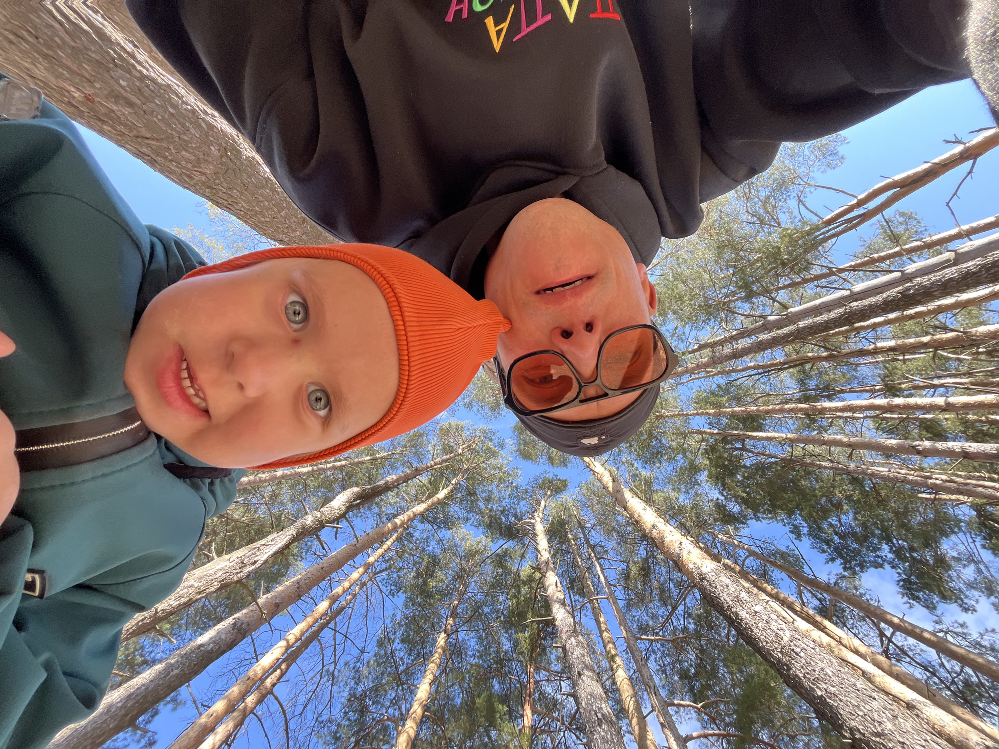 Captured from a low angle in what appears to be a forest in Chaikovsky, Russia, the image features two individuals - a child and an adult - with their faces close to the camera.
The child, wearing a distinctive orange beanie, is on the left side of the frame. Their face is angled towards the viewer, and a small portion of a green jacket with a strap is visible on their left.
On the right, an adult is wearing sunglasses and a black hoodie with multi-colored text. Above and behind them, numerous tall trees stretch upwards, their thin trunks reaching towards a bright blue sky with sparse clouds. The perspective emphasizes the height of the trees, creating a forest canopy effect.