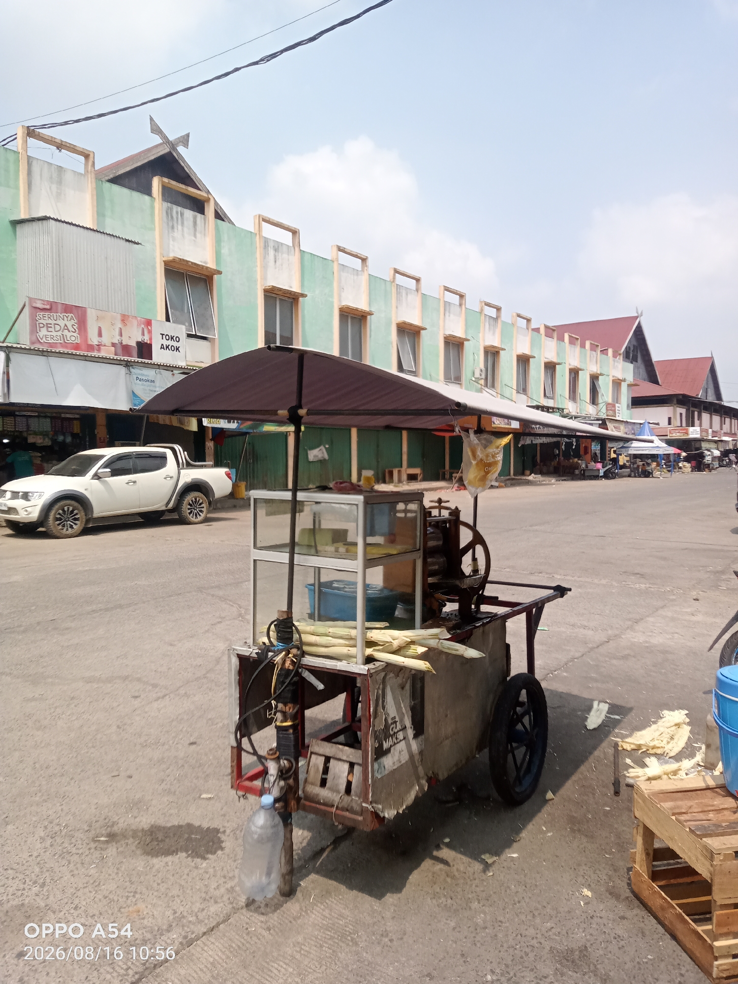 The image depicts a street scene in the city of Jambi, Indonesia, likely a market area. In the foreground, a sugarcane juice cart with a manual press is parked on an asphalt road. The cart is covered by a dark umbrella and features a glass display case holding what appears to be sugarcanes. Sugarcane stalks are also visible on the cart and scattered on the ground nearby. A plastic bottle is hanging from the cart. In the middle ground, a white pickup truck is parked on the side of the road. Beyond the truck, there are several buildings with green and beige facades, some with multiple stories and prominent rooflines. These buildings appear to house shops or stalls, with awnings and displays visible. The sky is bright and mostly clear with some clouds. The overall impression is that of a bustling outdoor commercial area on a sunny day.