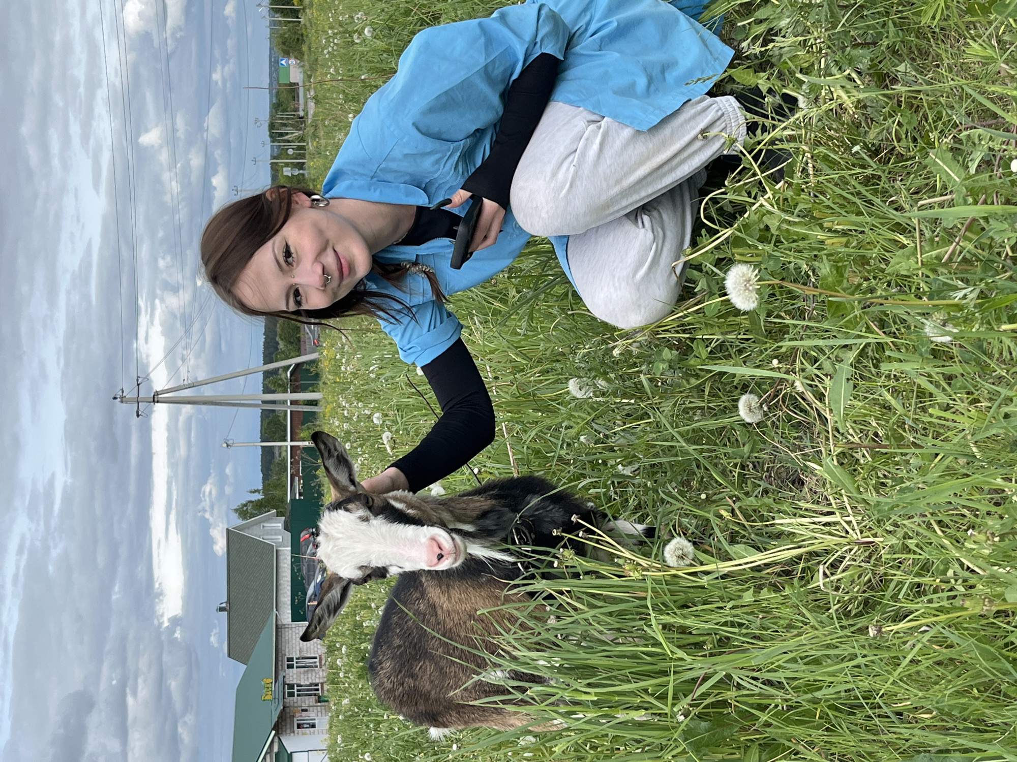 The image shows a woman kneeling in a grassy field with a goat. The woman is wearing a blue top, black undershirt, and grey pants, and has brown hair. She is smiling and looking at the camera, her left hand resting on the goat. The goat is brown, black, and white, and is standing in the grass. The background shows a building, power lines, and a cloudy sky. The location is Ludorvay, Russia, identified by the building in the background.