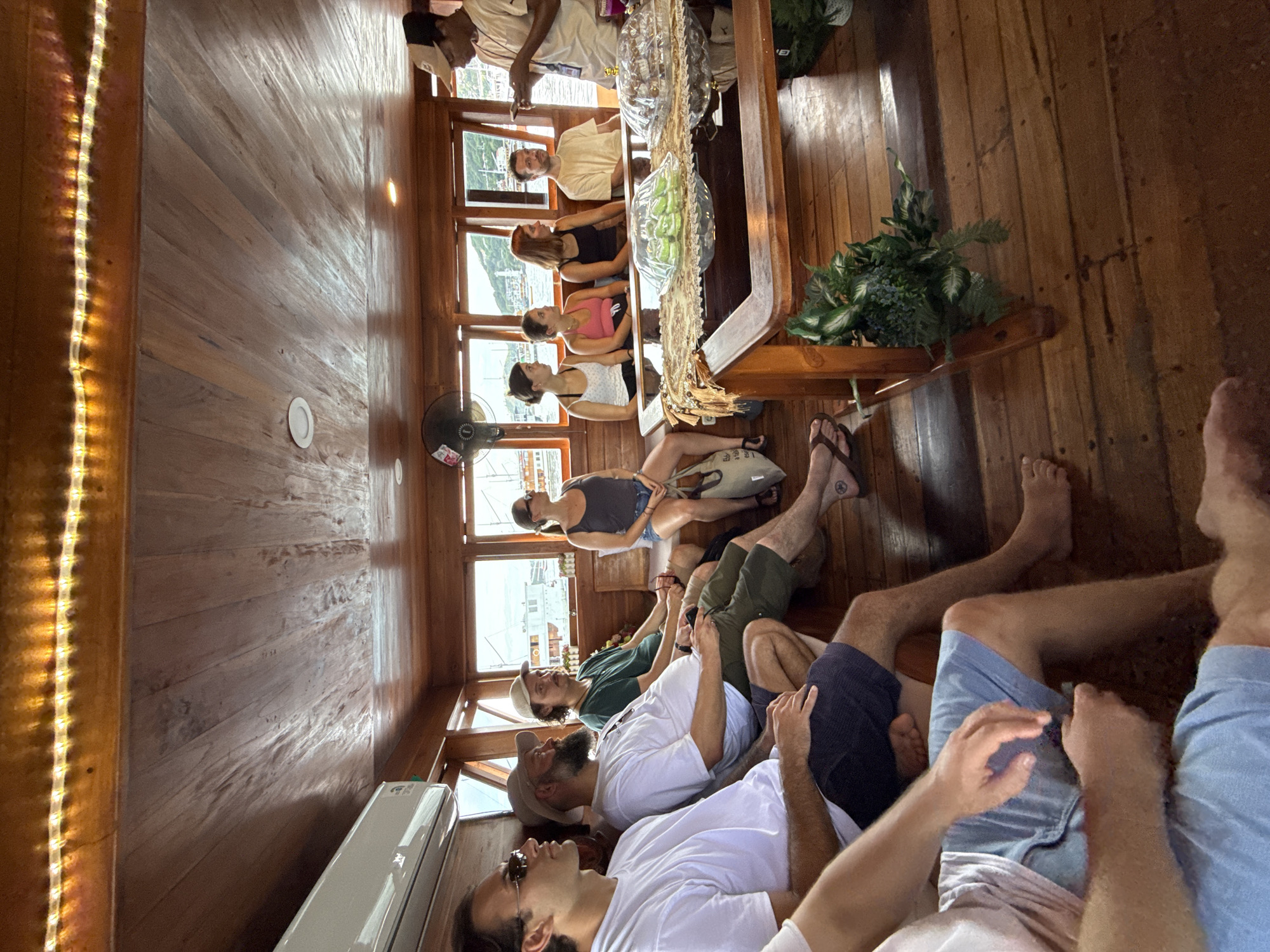 The photograph depicts the interior of a wooden boat in Labuan Bajo, Indonesia. There are ten individuals visible in the frame. Six men are reclining in the foreground, wearing casual clothing such as t-shirts and shorts. Four women are seated at a table near the center of the frame, listening to a man who appears to be a tour guide. The tour guide is standing, wearing a light-colored shirt and holding what appears to be a mobile device. The table is adorned with plants and decorative objects. Through the boat's windows, one can see a view of the sea and the town. A fan is mounted on the wall, and an air conditioning unit is near the bottom left.