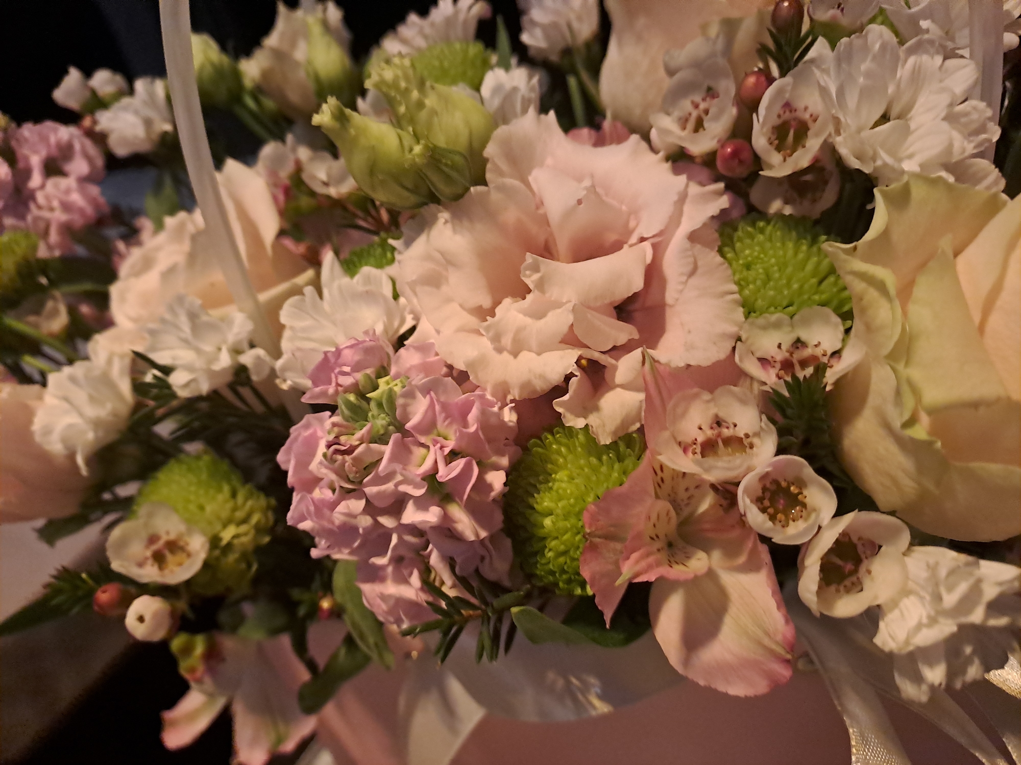 This is a close-up, eye-level, indoor photograph of a floral arrangement. The arrangement contains a variety of flowers in shades of pink, white, and green, including large, pale pink roses or peonies, smaller white flowers with visible stamens, and bright green, spherical flowers. There are also some closed green buds and delicate pink flowers with darker centers. The flowers are densely packed and appear to be in a light-colored container, possibly a basket, with ribbons visible. The background is dark and out of focus, suggesting artificial lighting. There are no people or animals in the image. The setting is domestic or a florist shop. There is no indication of the location being in Krasnyi Ogorok, Russia.