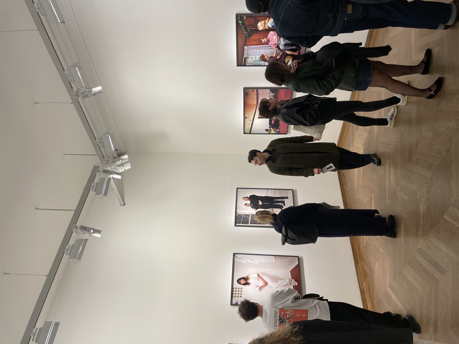 People are in an art gallery, viewing framed photographs on a white wall. The pictures range from portraits to interior scenes. Viewers are dressed in casual attire, wearing jackets, dresses, and jeans. A track lighting system is installed on the ceiling. The floor is a light-colored wood.
