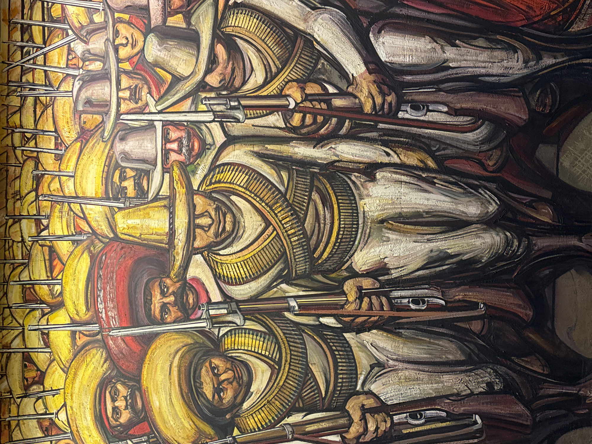 The image is a section of a mural in Mexico City, Mexico. It depicts a large group of Mexican revolutionaries wearing sombreros and bandoliers, holding rifles with bayonets attached. They are standing shoulder-to-shoulder in rows. The figures are rendered in a stylized, possibly expressionistic manner. The colors are predominantly warm, with yellows, browns, and ochres.