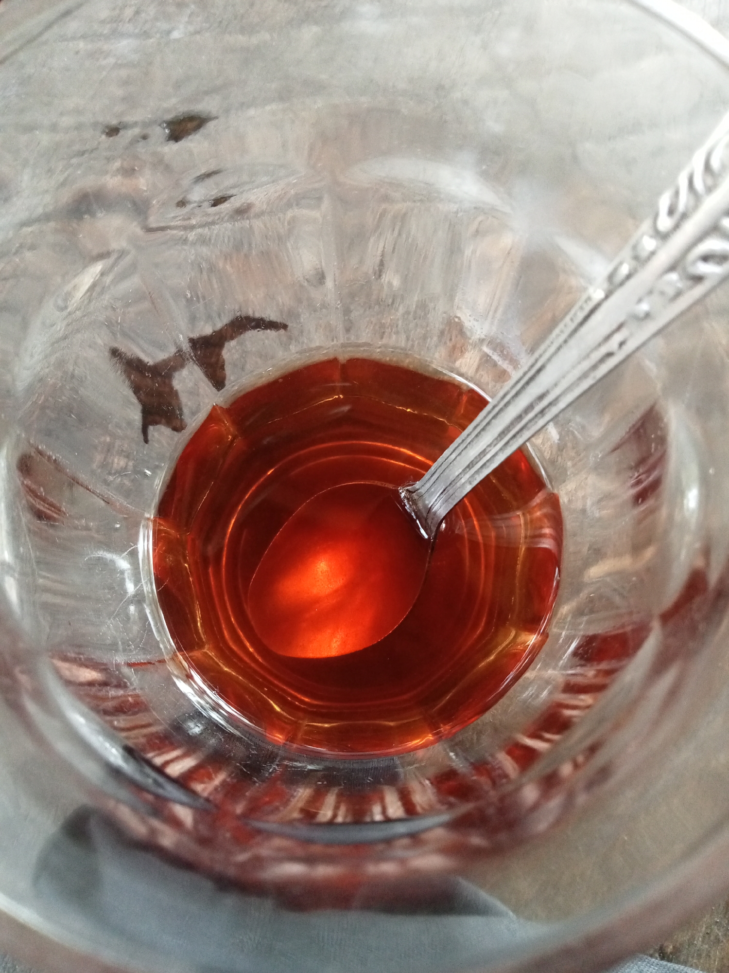 An overhead shot shows a glass with a reddish-brown liquid and a spoon inside, in Matangsijuek Timur, Indonesia. The glass has a textured, ribbed surface and appears to be partially filled with the liquid. The spoon, made of a shiny metal, rests within the liquid, with its handle extending out of the glass. The liquid is translucent and reflects light, creating a bright highlight on the spoon and the surface of the liquid. The background is blurred, but appears to be a wooden surface.