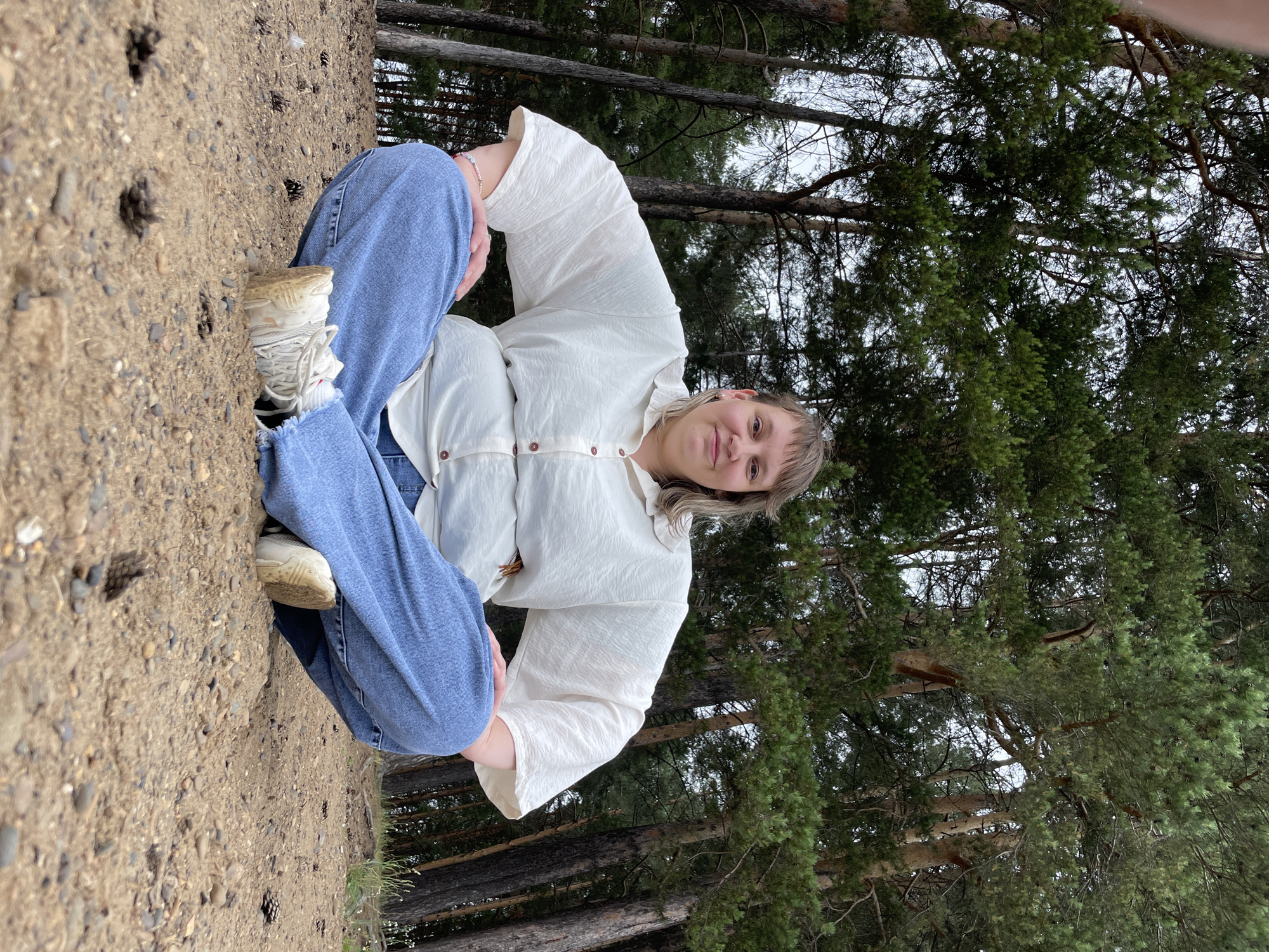 A medium shot captures a person sitting cross-legged on sandy ground in a forest. The person has light skin, light brown hair styled in layers, and is smiling at the camera. They are wearing a loose, off-white button-up shirt with wide sleeves and light blue denim jeans. Their feet are clad in white sneakers. The ground is covered in sand, small pebbles, and pinecones. Behind the person, a dense forest of tall pine trees with green needles forms the backdrop. The sky is overcast, and the overall lighting is subdued. The scene is likely in the city of Chaikovsky, Russia, given the prevalence of pine forests in the region.
