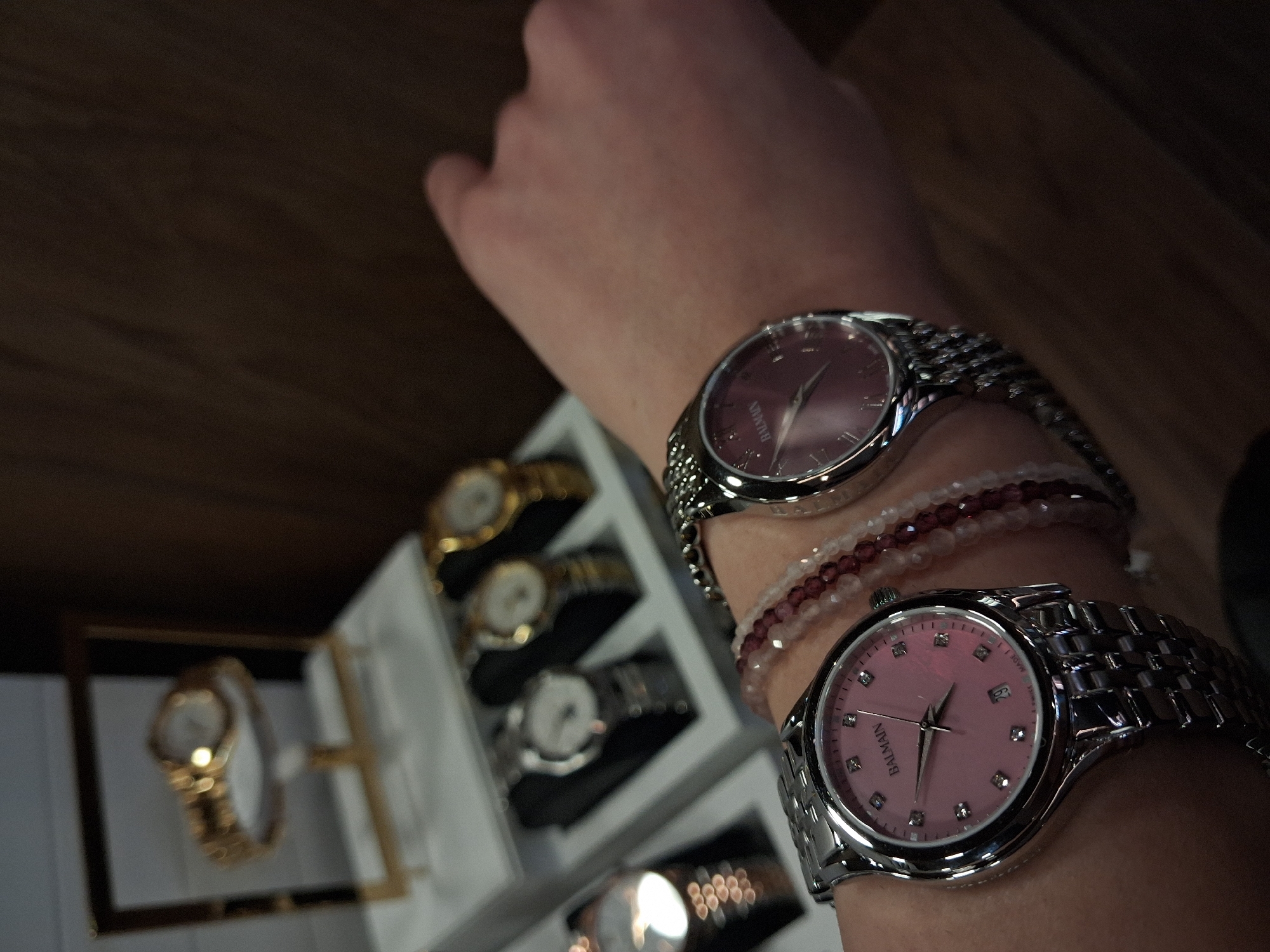 The image shows a person's arm wearing two Balmain watches and a beaded bracelet. The watches are silver with pink and purple faces. The bracelet is a mix of white and red beads. Behind the arm and watches is a watch case with several other watches displayed, including gold and silver ones. The location is not specified in the image. There is no reference to Varna, Bulgaria, within the image.