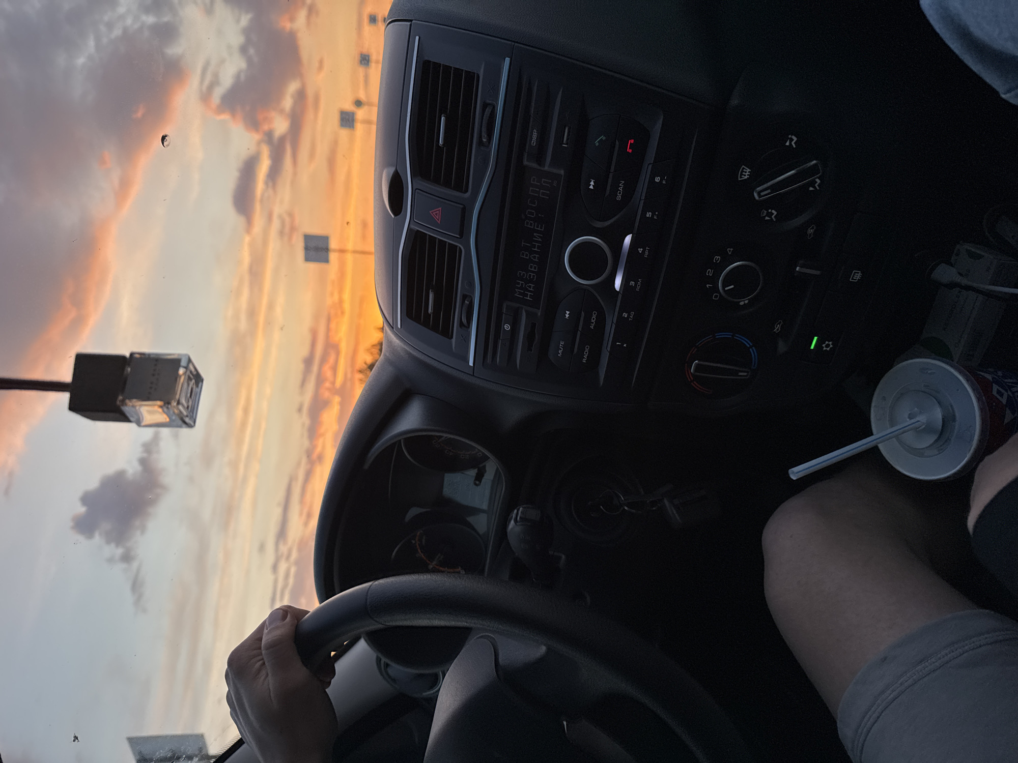 The image captures a first-person perspective from inside a vehicle, looking towards a beautiful sunset or sunrise sky. A person's hand is visible on the bottom part of the steering wheel, indicating they are driving or a front-seat passenger. Part of their leg, wearing gray shorts, is also visible on the right, next to a white and red beverage cup with a straw.
The car's interior features a dashboard and center console. The stereo system has a display showing Cyrillic text: "МУЗ ВТ ВОСПР НАЗВАНИЕ: пл", which translates to "Music BT Playback Name: pl" (likely 'playlist' or 'play'). Various buttons are visible around the stereo, including "MUTE", "RADIO", "AUDIO", "SCAN", and numbered presets (1-6). Below the stereo are climate controls with dials for fan speed, temperature, and air direction. Car keys with a keyring are seen hanging from the ignition on the lower left of the console. An air freshener hangs from the rearview mirror, visible at the top left of the windshield.
Outside the car, the sky is filled with warm, vibrant colors of orange, pink, and purple, indicating either dawn or dusk. Clouds are scattered across the sky. There appear to be reflections or structures of what might be streetlights or poles in the distance. The windshield has some dust or small specks visible, adding to the authenticity of the roadside view in Nagornaya, Russia, as suggested by the provided context and the Cyrillic text on the car stereo. The overall mood is calm and relaxed, perhaps a moment of appreciation for the scenic view during a drive.