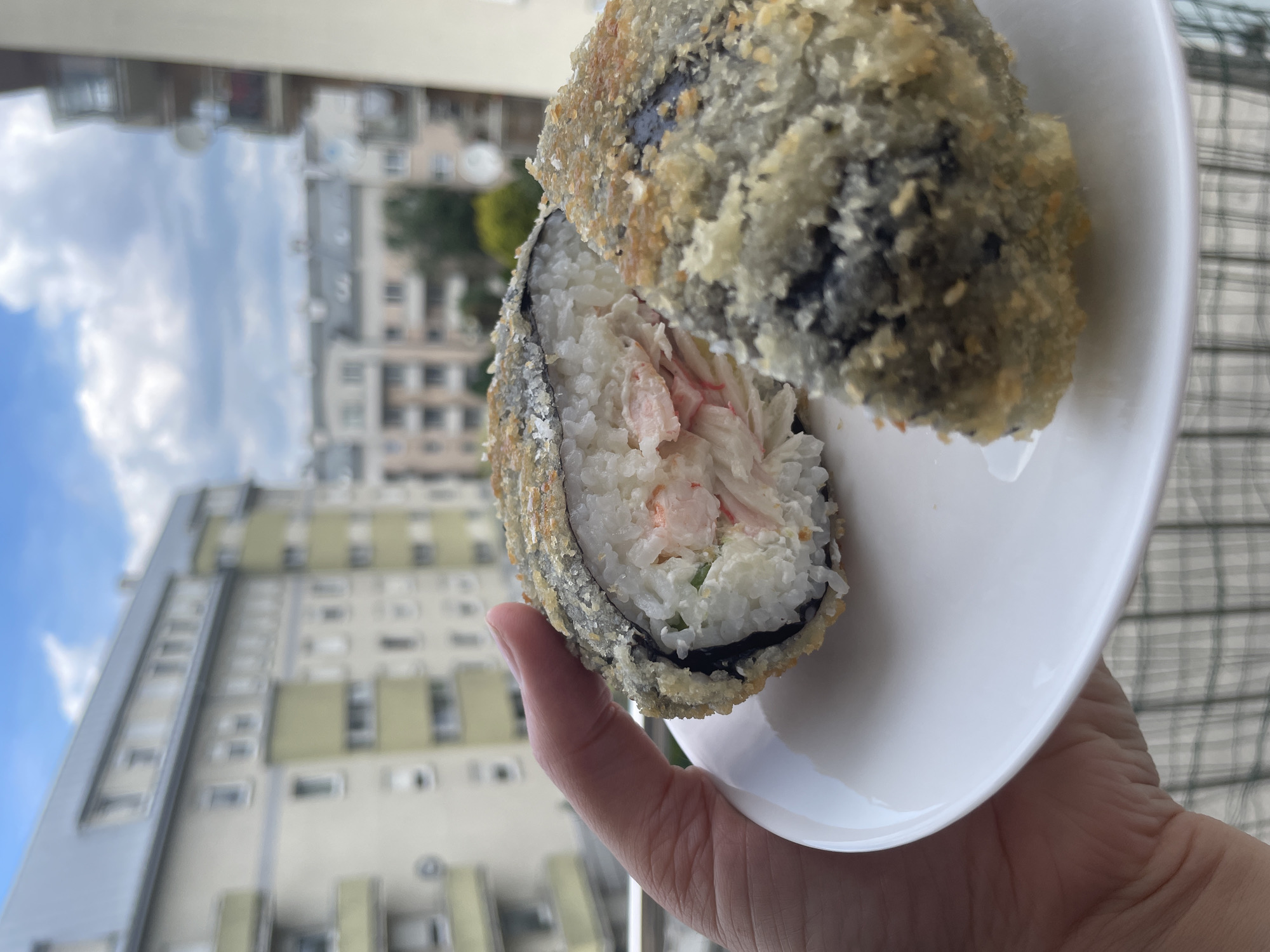 A person's hand holds a white plate with a deep-fried sushi roll on it. The sushi roll is cut in half, revealing white rice and pink crab meat filling. The outer layer of the sushi is golden brown and crispy, with visible bits of seaweed. In the background, blurred buildings with balconies and windows are visible under a cloudy sky.
