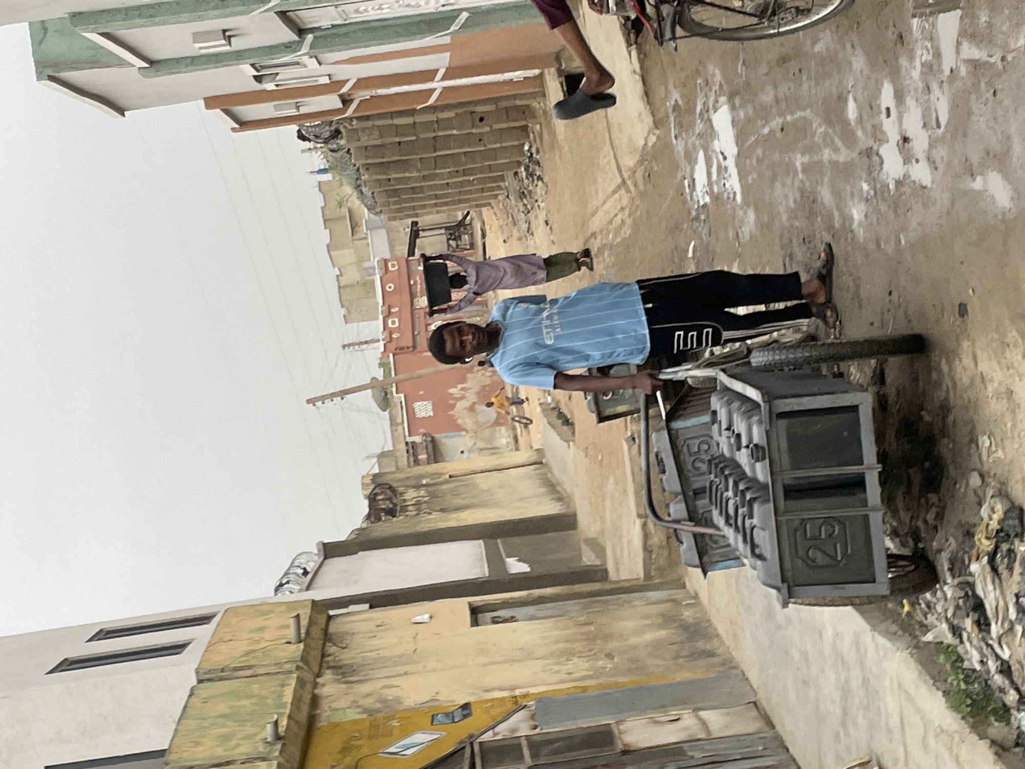 The image shows a man pulling a heavy, metal cart down a dirt street in a city. The cart is gray and appears to have a number of compartments with the number 25 visible. Buildings line the street, constructed of beige and brown materials, some with visible damage. Overhead power lines crisscross the sky. Another individual stands further down the street holding a large, dark, rectangular object. A parked bicycle is visible in the upper portion of the frame, along with another person's legs. The scene appears to be in a residential neighborhood, and there's litter along the edge of the street. The location appears to be the city of Unknown, Unknown.