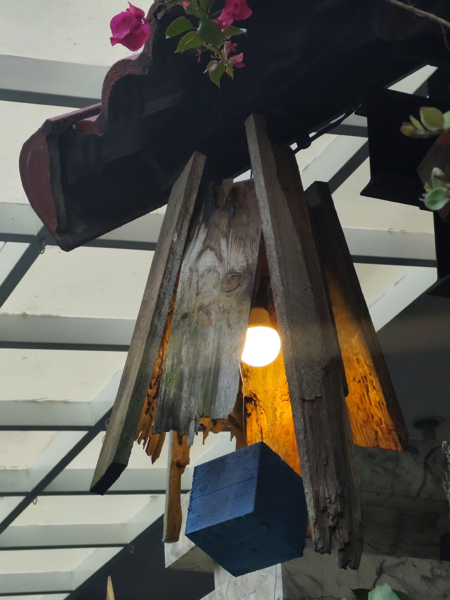 The image shows a rustic, handmade light fixture made of weathered wood. The fixture is suspended from a dark, roof-like structure adorned with a few pink flowers and green leaves. The wooden elements are rough and uneven, with visible grain and signs of age. A single, illuminated lightbulb hangs within the wooden frame, casting a warm glow. Below the light fixture, a dark blue, cubical object is also suspended. The background reveals a structure with white and grey, angled panels, suggesting an outdoor or covered area, possibly a patio or a building's exterior. The overall impression is of an artfully constructed, unconventional lighting element. The image is taken from a low angle, looking upwards. There are no people visible in the scene. This fixture is located at the exterior of a cafe named "Tiệm Cà Phê Tùng" (Tung Cafe) in Dalat, Vietnam.