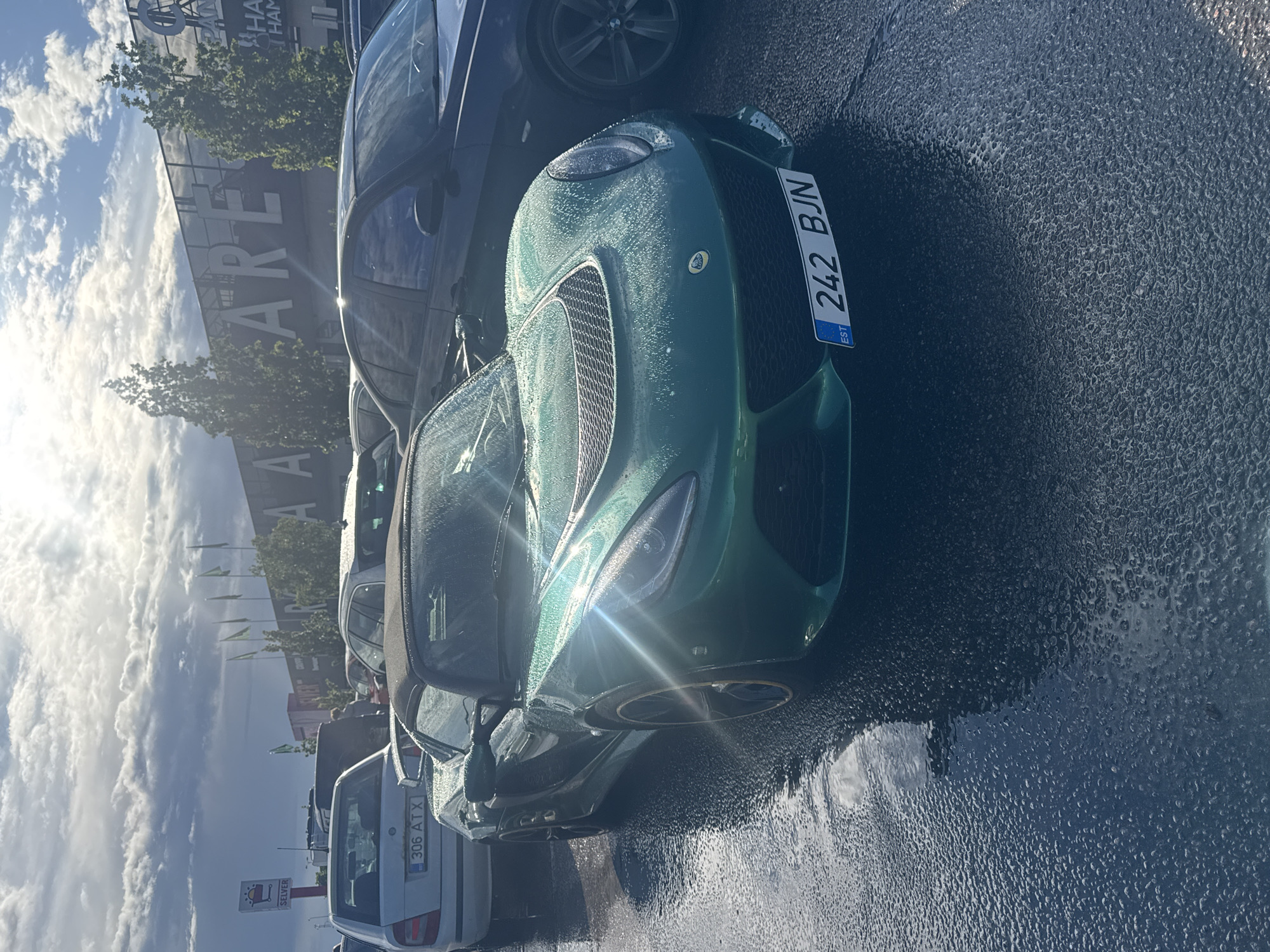 The image shows a green Lotus sports car parked on a wet asphalt surface. The car has a license plate that reads "242 BJN EST". Behind the sports car, other vehicles are parked, including a dark-colored sedan and a silver hatchback. In the background, a large building with large white lettering that appears to say "ARENA" is visible. Several green flags are also flying in front of the building. The sky is cloudy with sunlight breaking through. The scene suggests a parking lot in Tallinn, Estonia, likely near an arena or large commercial building.