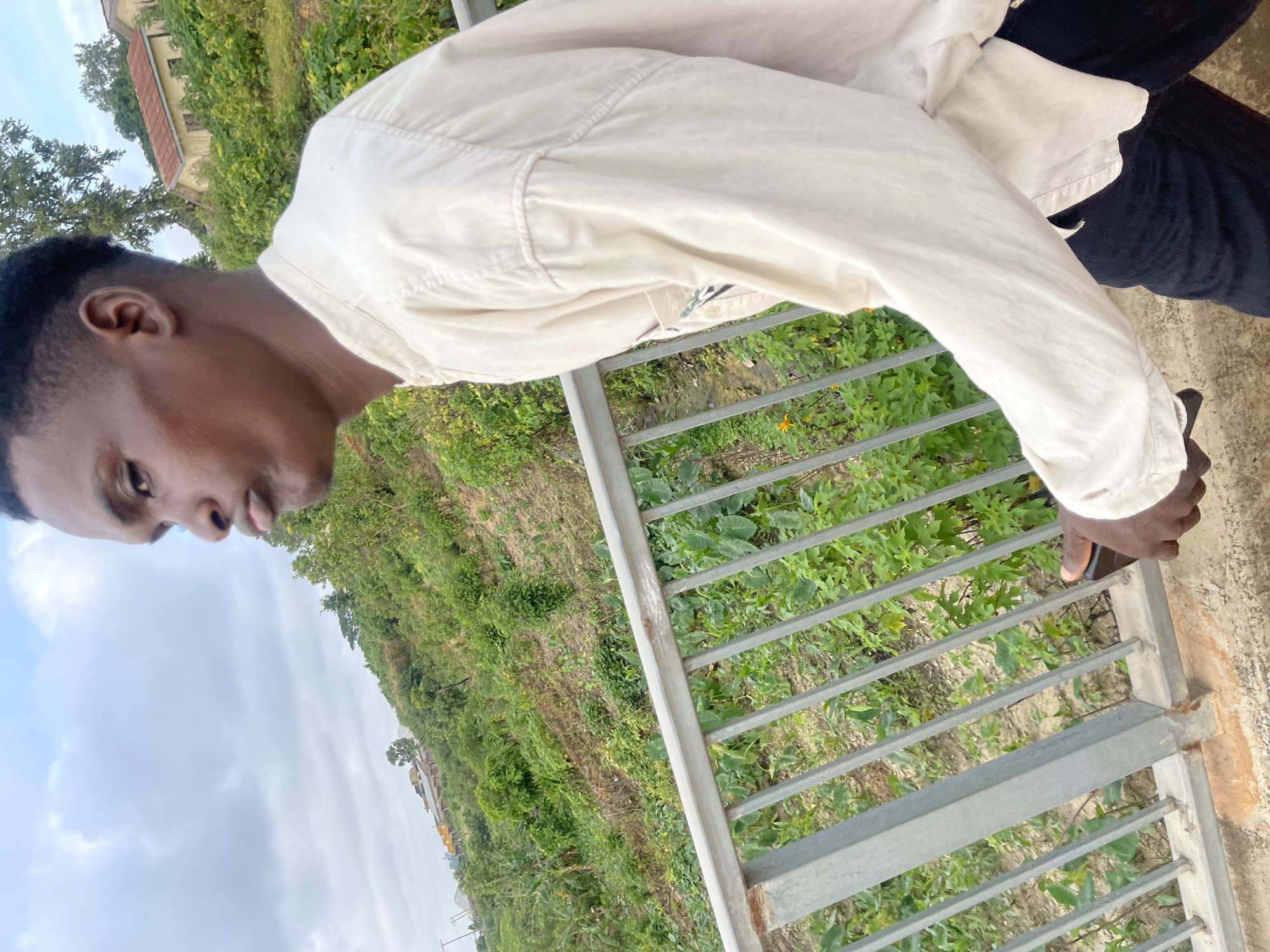 A young Black man stands on a walkway, his body angled away from the camera. He wears a light-colored, long-sleeved collared shirt and dark pants. His left hand is visible, holding a dark rectangular object, possibly a phone. The man is looking off to his right, his lips pursed.
Behind him, a metal railing separates the walkway from a steep, green hillside dotted with various plants and some scattered buildings. The hillside appears to be a rural or semi-rural area. The sky above is overcast. The scene is outdoors. There is no indication of the specific location being Rafin Par, Nigeria.