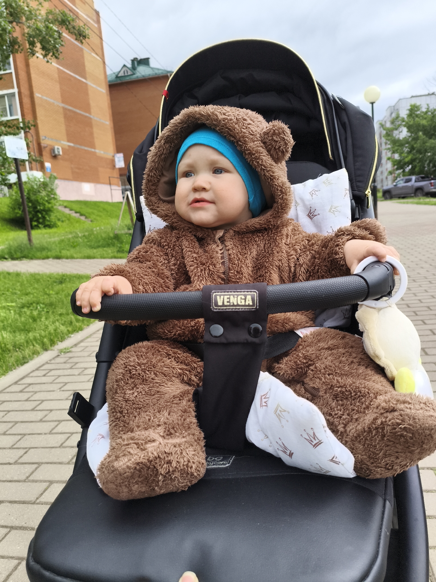 An outdoor, slightly overcast scene in Chaikovsky, Russia, features an infant in a black stroller on a paved brick sidewalk. The infant, appearing to be approximately 6-12 months old, is dressed in a brown, plush, full-body jumpsuit resembling a bear, complete with small ear appendages on the hood. A light blue cap is visible underneath the hood. The infant's hands grasp a black textured bar positioned in front of them within the stroller. A small, light yellow plush toy is attached to the right side of this bar. The stroller's chest harness is visible, secured over a white fabric insert patterned with brown and gold crowns. The stroller itself bears a "VENGA" brand label on the central bar.
In the background to the left, a multi-story brick building with orange and brown facades is visible, partially obscured by green foliage. Further back and slightly to the right of the central building, another structure with a dark green roof is discernible. To the right of the stroller, a tall, spherical yellow streetlight stands beside the sidewalk. Beyond the streetlight, green grass and additional trees line a paved road where a grey pickup truck is parked in the distance. The sky above is cloudy.