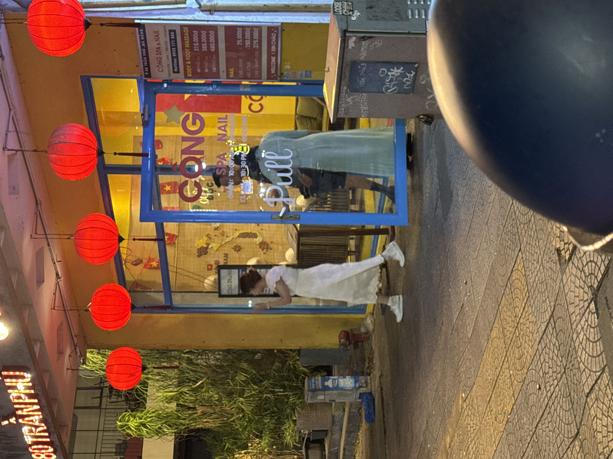 The image depicts a street scene at night in Da Nang, Vietnam, likely on Tran Phu street, indicated by the neon sign "80 TRAN PHU". Red paper lanterns are suspended overhead, illuminating the entrance of a spa and nail salon named "CÔNG SPA & NAIL". A woman in a white dress and white sneakers is walking away from the camera, towards the spa. Inside the salon, a man wearing dark clothing and a woman in a light green coat are visible through the blue-framed glass door, suggesting an interaction or waiting. The pavement is paved with patterned tiles. Signs inside the salon display prices for services like "BODY & FOOT MASSAGE" and "NAIL". The overall ambiance is that of a commercial street at night, with a focus on the entrance and the people associated with the establishment.