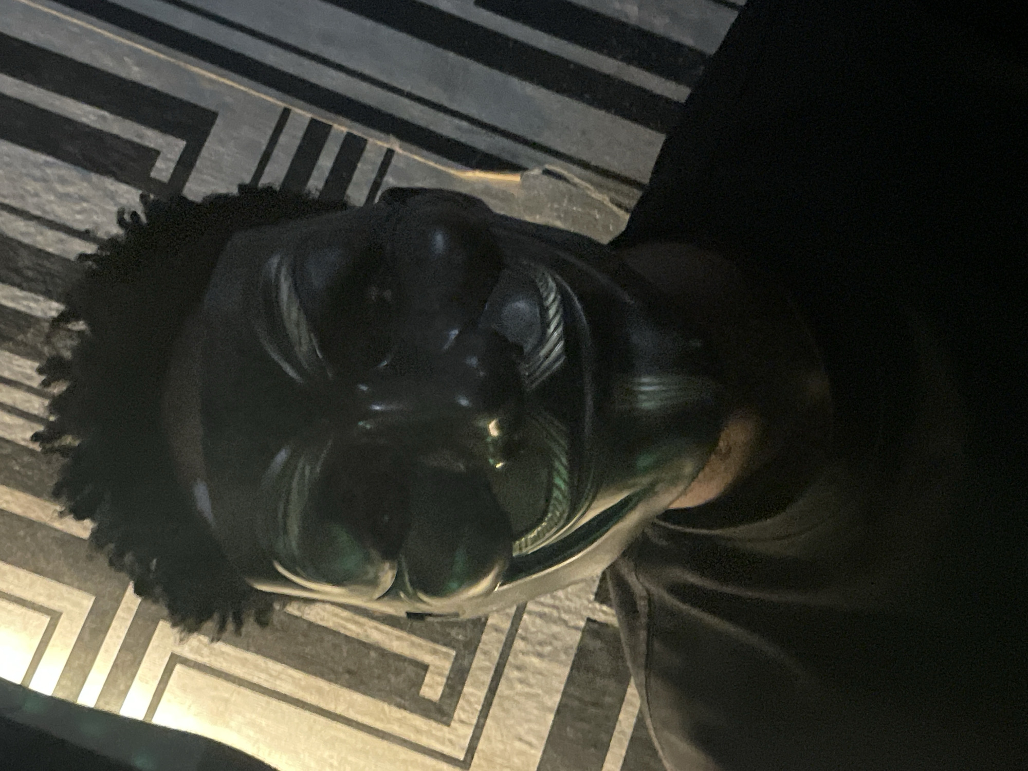 A person with dark hair is wearing a black mask with a shiny finish. The mask covers the entire face, with visible contours suggesting the shape of facial features underneath. The person is wearing a black garment. The background consists of a surface with a geometric pattern of alternating black and light-colored lines. There is insufficient information to determine the location in Lokoja, Nigeria.