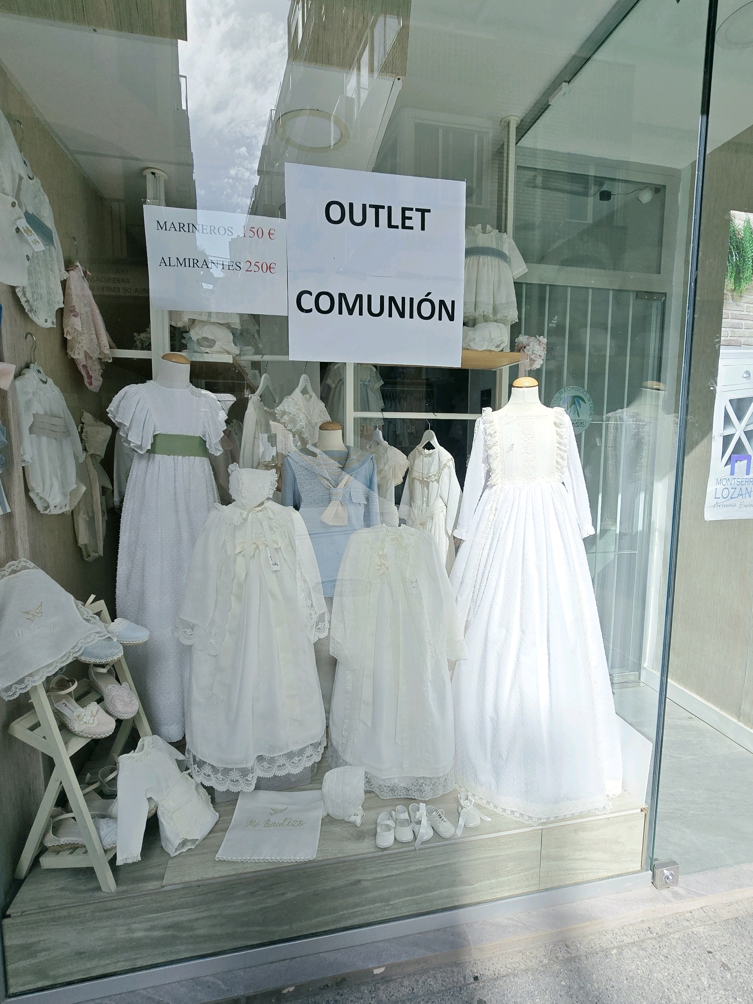 This image shows the storefront window of a children's clothing store, likely in Jaén, Spain, given the context provided in the crops. The display features various white and light-colored formal outfits for children, possibly for christenings or first communions, as indicated by the text "COMUNIÓN" visible on a sign. Several dresses, rompers, and accessories like bonnets and shoes are artfully arranged on mannequins and shelves. One large sign reads "OUTLET" and another indicates prices for "MARINEROS" (sailors) at 150€ and "ALMIRANTES" (admirals) at 250€. The window is made of clear glass, reflecting the sky and buildings outside. The store appears to be located on a street, with a sidewalk visible in the foreground. The overall impression is of a well-presented, upscale children's boutique.