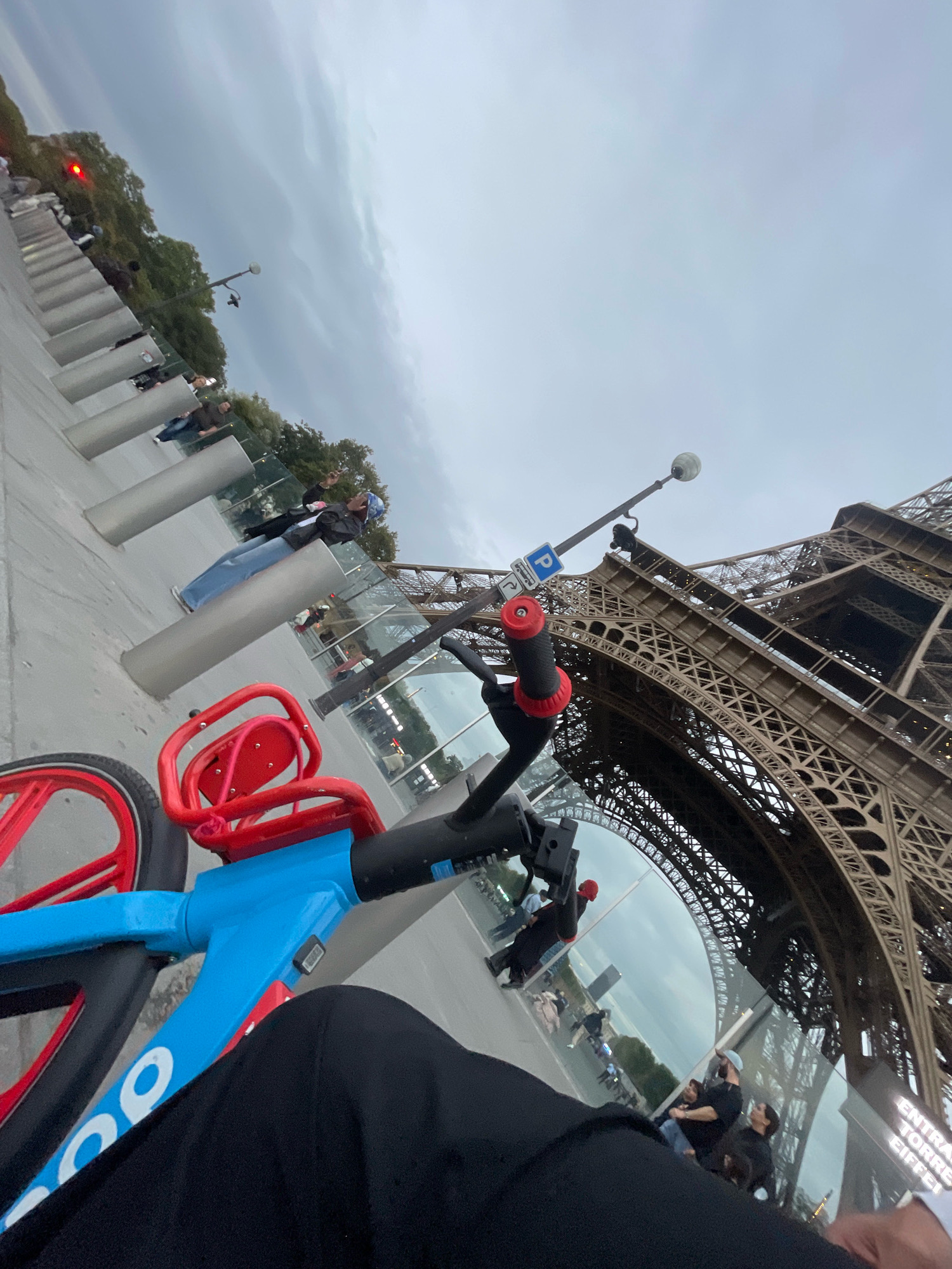 The image depicts a low-angle, wide shot of the Eiffel Tower in Paris, France. In the foreground, a blue and red electric scooter is partially visible, with the rider's black pants occupying the lower right corner of the frame. The Eiffel Tower dominates the background, its intricate iron lattice structure filling the right half of the image. A glass barrier separates the walkway from the area below, and several people can be seen walking and standing behind it. In the mid-ground, a line of cylindrical bollards runs along the edge of the walkway, and trees are visible in the distance. The sky is overcast with a soft, diffused light. A street lamp is positioned above and to the left of the Eiffel Tower, and a blue parking sign is attached to its pole.