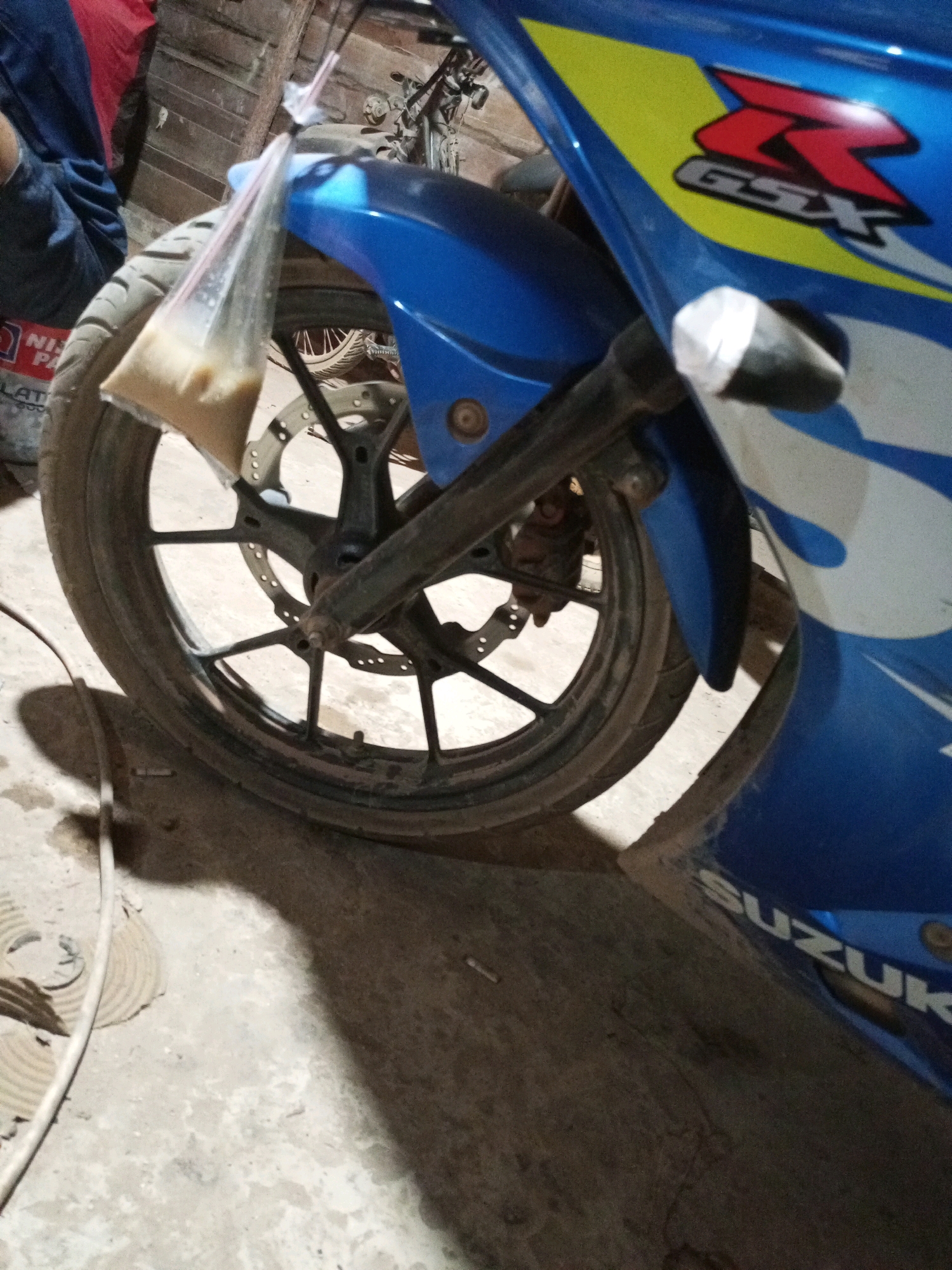 A close-up shot captures the front wheel of a blue Suzuki motorcycle, parked on a rough concrete floor. A clear plastic bag filled with a beige liquid is suspended from the handlebars by a straw. The motorcycle features the "GSX-R" logo in red and yellow on its fairing and "SUZUKI" lettering on its side. In the background, part of a person's leg in blue trousers and a red garment is visible, along with wooden planks and another motorcycle. The lighting suggests an indoor or shaded area.