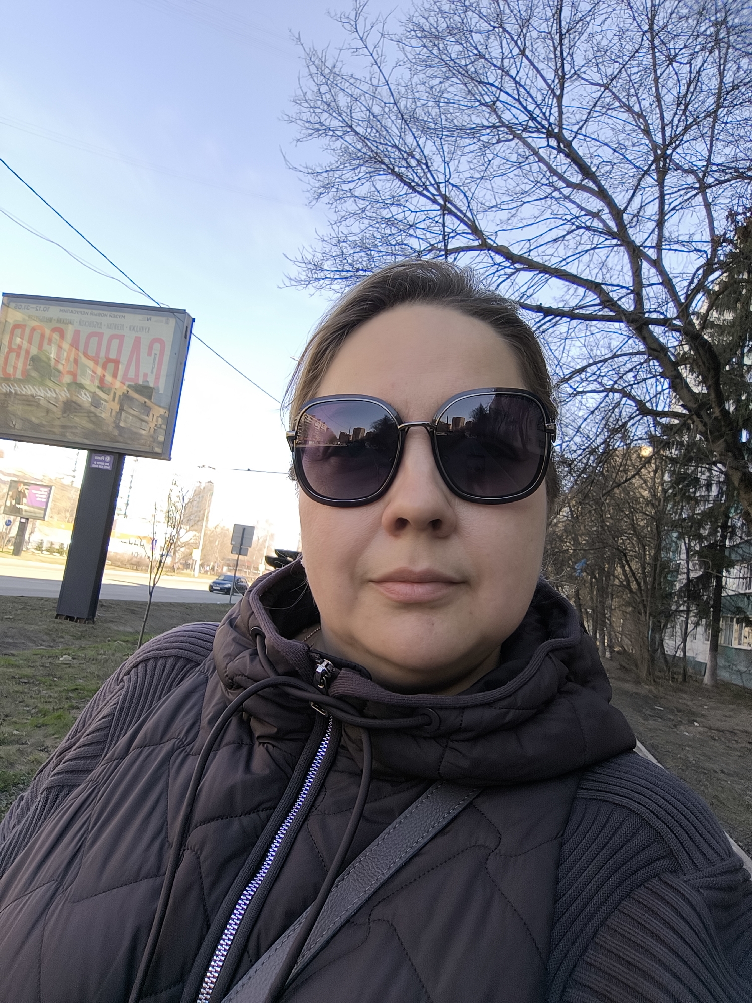 A woman wearing dark sunglasses and a brown quilted jacket with a grey shoulder strap stands outside. Behind her is a tree with bare branches against a blue sky. To the left, a billboard displays an advertisement in Russian that includes "САВВЕРСОВ". Buildings and trees are also visible in the background. Based on the "САВВЕРСОВ" on the billboard, which is a business in Khimki, Russia, and the overall appearance of the surroundings, the location is likely Khimki, Russia.