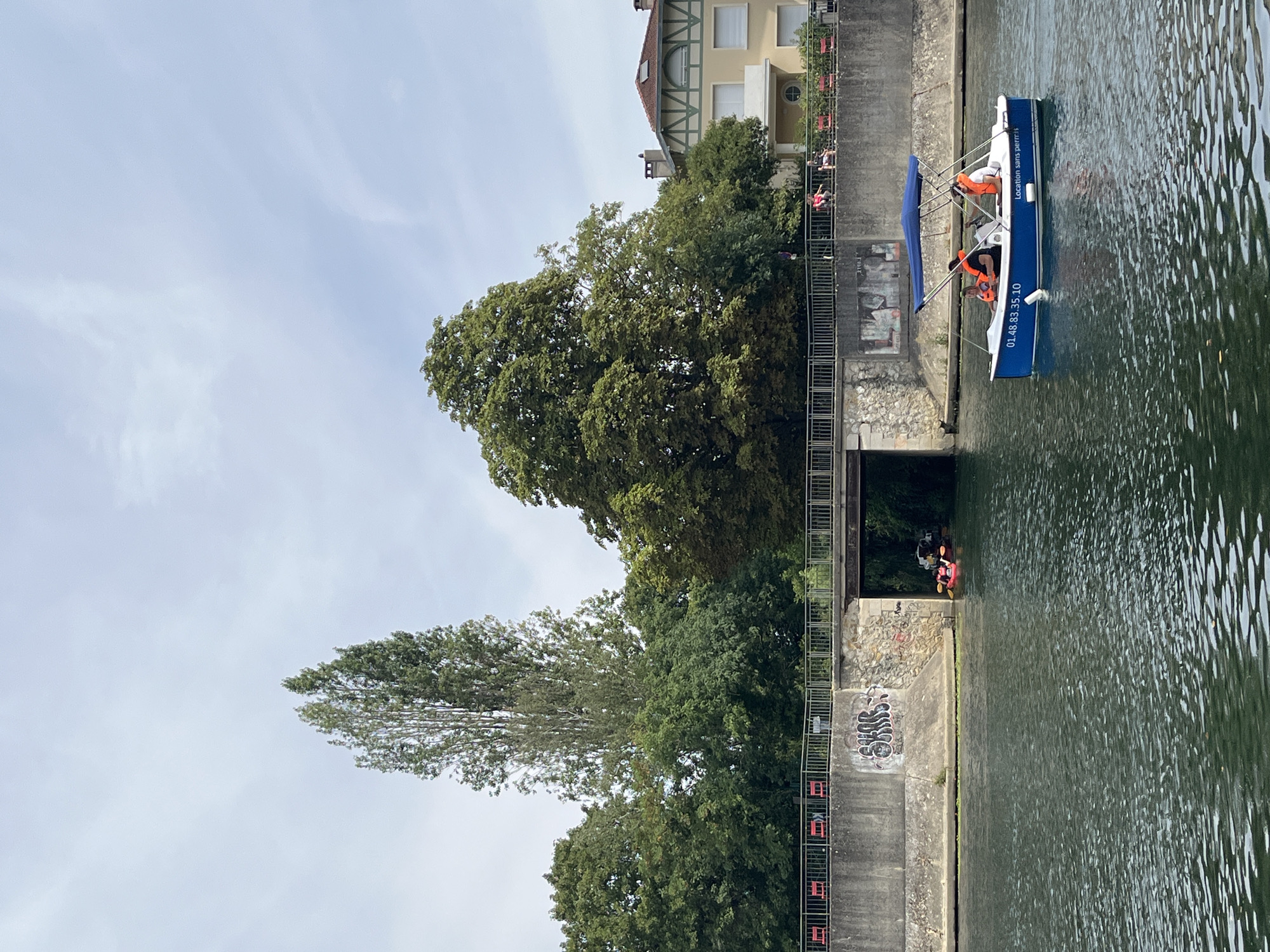 In the city of Joinville-le-Pont, France, on a body of water, a small blue motorboat with a blue canopy is moving from right to left. Three individuals are aboard the boat, two wearing orange life vests. On the left side of the image, two people in a red kayak are visible near a bridge. The water has a textured, rippling surface. Behind the water, a concrete retaining wall with graffiti runs horizontally. A bridge spans the water, supported by stone and concrete structures, with people and red chairs visible on the walkway above. Lush green trees form a dense backdrop, with a tall, slender tree on the left and a large, mature tree on the right. A light-colored building with a red-tiled roof and green trim is partially visible behind the trees on the far right. The sky is overcast with scattered clouds. The boat displays the text "01.48.83.35.10" and "Location sans permis" on its side.