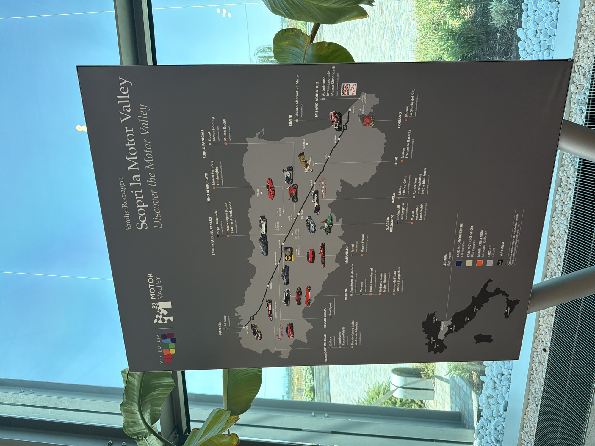The image shows a large map poster titled "Emilia-Romagna Scopri la Motor Valley / Discover the Motor Valley." The map displays the Emilia-Romagna region of Italy with various automotive-related locations marked by icons and text. The poster is presented in a modern, well-lit indoor space with large windows and green foliage visible in the background, suggesting a museum or exhibition hall. The map itself is dark gray with white text and colorful icons representing car manufacturers, museums, and circuits. The bottom section of the poster includes a legend that color-codes different types of attractions: blue for car producers, orange for bike producers, red for museums and collections, and gray for circuits. A small inset map of Italy is also present. The lighting suggests it is daytime. There are no people visible in the image. The location is identified as being in Sant'Anna, Italy, based on a previous context not present in the image.