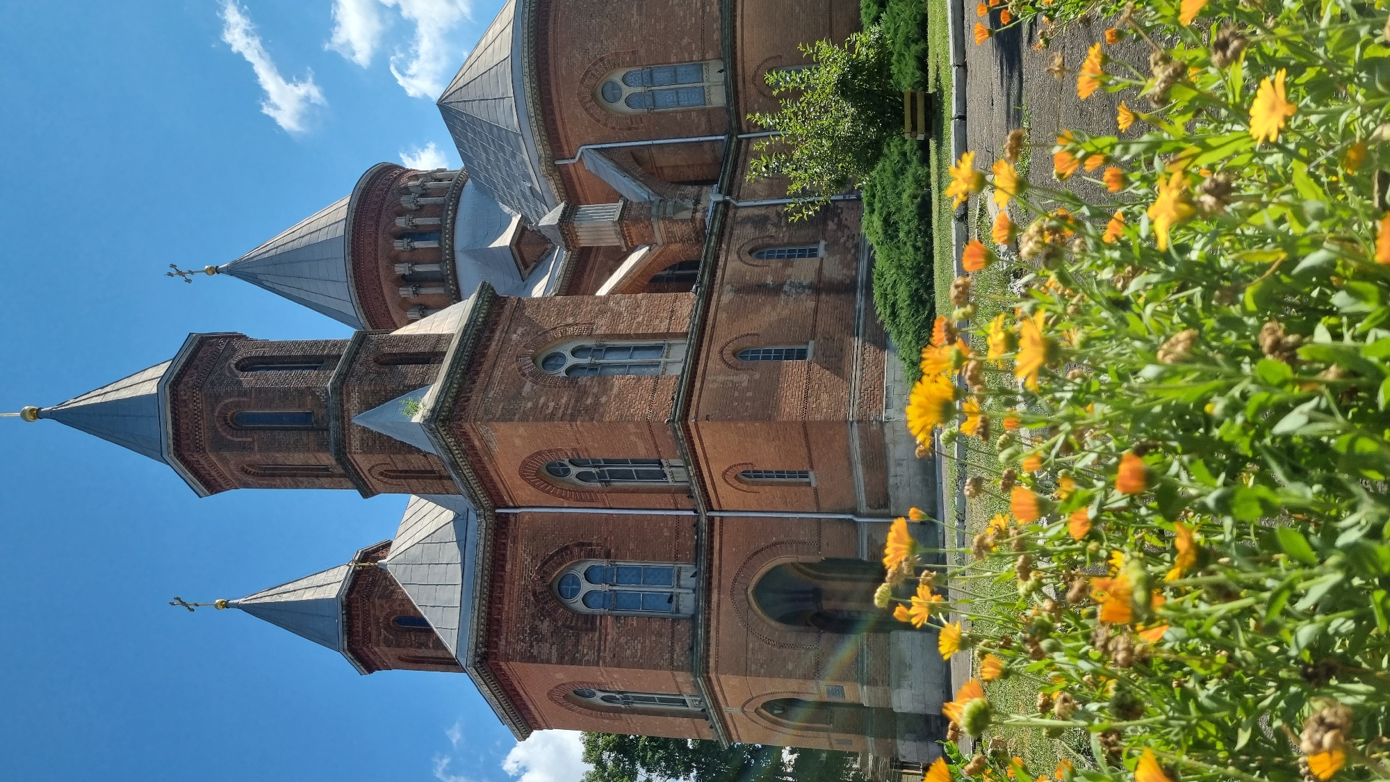 This is a vertical, outdoor, eye-level shot of the Chernivtsi National University main building, also known as the former Residence of Bukovinian and Dalmatian Metropolitans, located in Chernivtsi, Ukraine. The structure is made of red brick and features multiple towers with conical, grey roofs topped with crosses. The windows are arched with Gothic architectural elements. In the foreground, a dense patch of bright orange and yellow calendula flowers blooms in the sun. The sky is bright blue with scattered white clouds. The overall lighting suggests it is a sunny day.