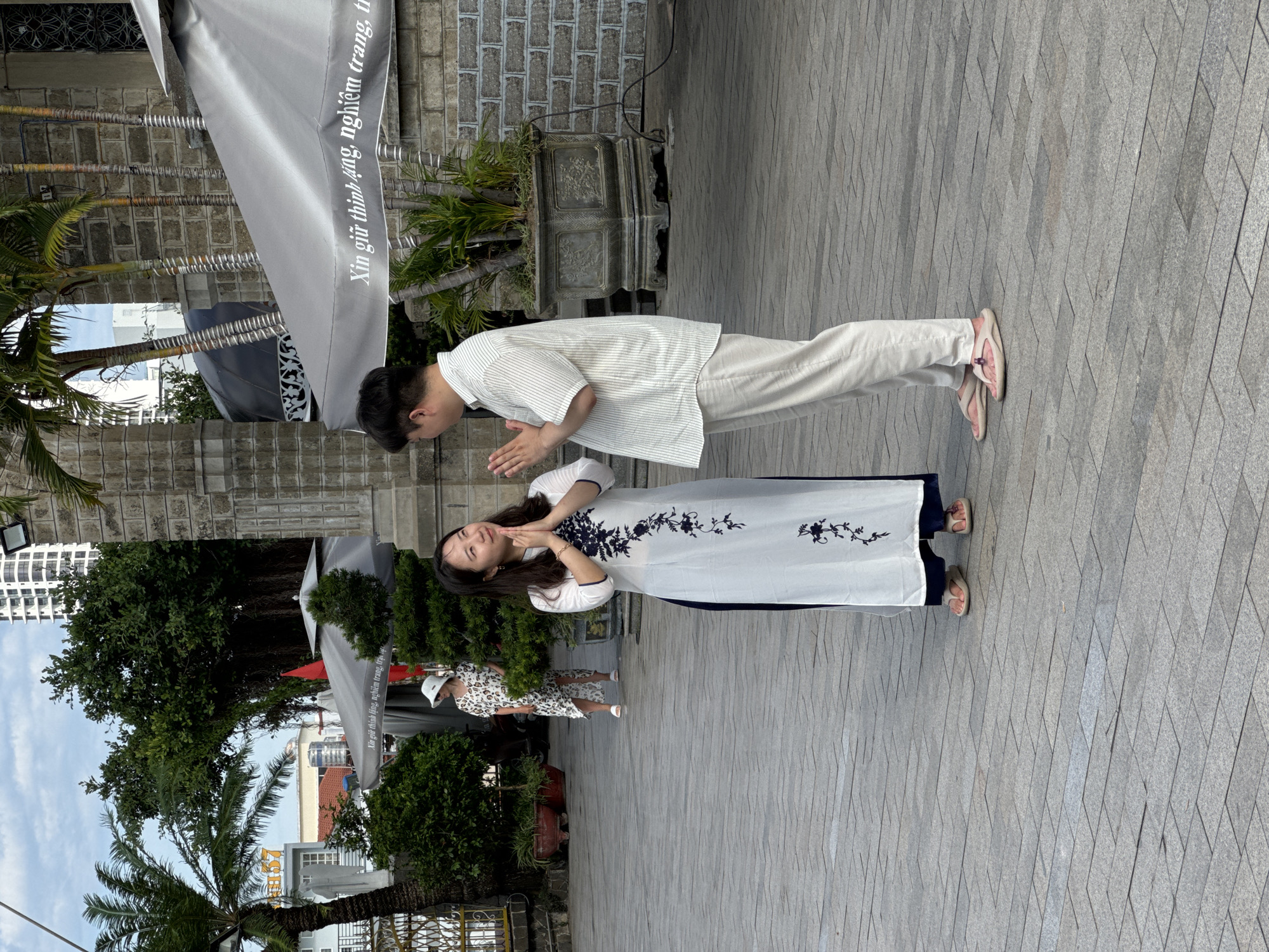 The image captures a scene in Nha Trang, Vietnam, likely near a historical or religious site given the architectural elements visible. In the foreground, a woman in a white traditional Vietnamese dress, a long tunic with dark blue floral embroidery, stands with her hands pressed together in a prayer-like gesture. She is looking upwards with a gentle expression. Facing her is a man in a casual, light-colored striped shirt and trousers, also with his hands clasped in a similar gesture. The two individuals are positioned on a paved courtyard.
In the background, another woman in a patterned dress and a white hat is visible, walking away from the foreground subjects. Behind the main subjects, parts of a building constructed with stone bricks are seen, along with palm trees and lush green foliage. Large gray umbrellas, common in outdoor dining areas or event spaces, are also present. The setting appears to be an open public area, possibly adjacent to a church or a temple, given the architectural style and the presence of umbrellas that might shade an outdoor cafe or waiting area. The sky is partly cloudy, suggesting it is either early morning or late afternoon. The overall atmosphere is peaceful and contemplative.