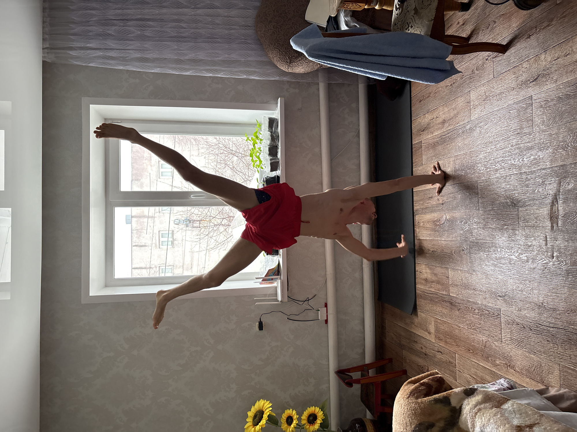A Caucasian man in red shorts is performing a yoga pose in an apartment in Ulyanovsk, Russia. He is balancing on one hand on a black yoga mat on a wooden floor, with his legs spread in a wide split. The view from the window shows an apartment building and bare trees. Other objects include a sunflower, plants, a curtain, and a chair.