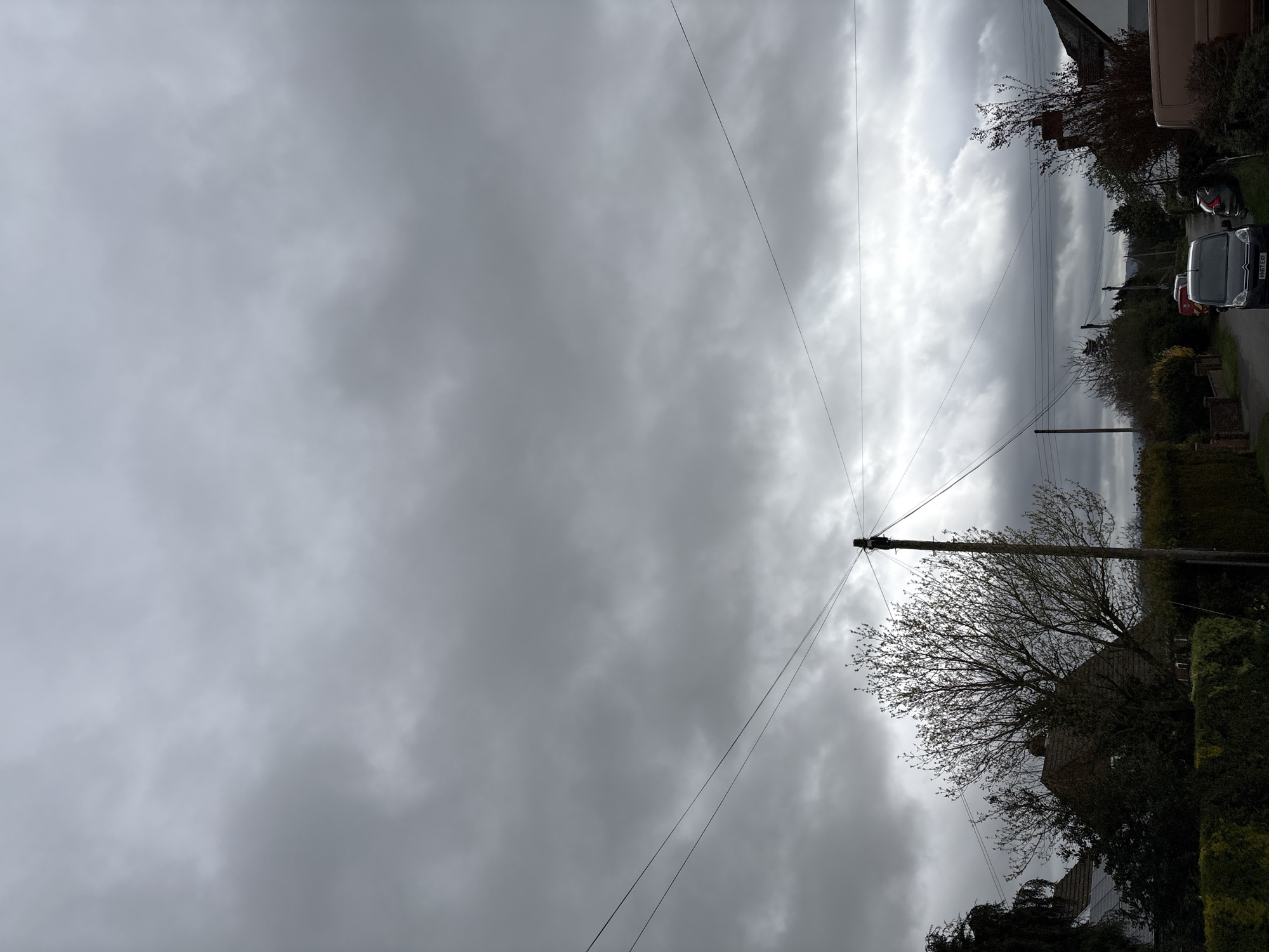 The image depicts a street scene in Longden, United Kingdom, under an overcast sky with dense cloud cover. Power lines run across the upper part of the image, converging towards a utility pole on the right side. Buildings and trees line the right side of the street. Several parked vehicles are visible, including a white van. The road appears to be a narrow residential street.