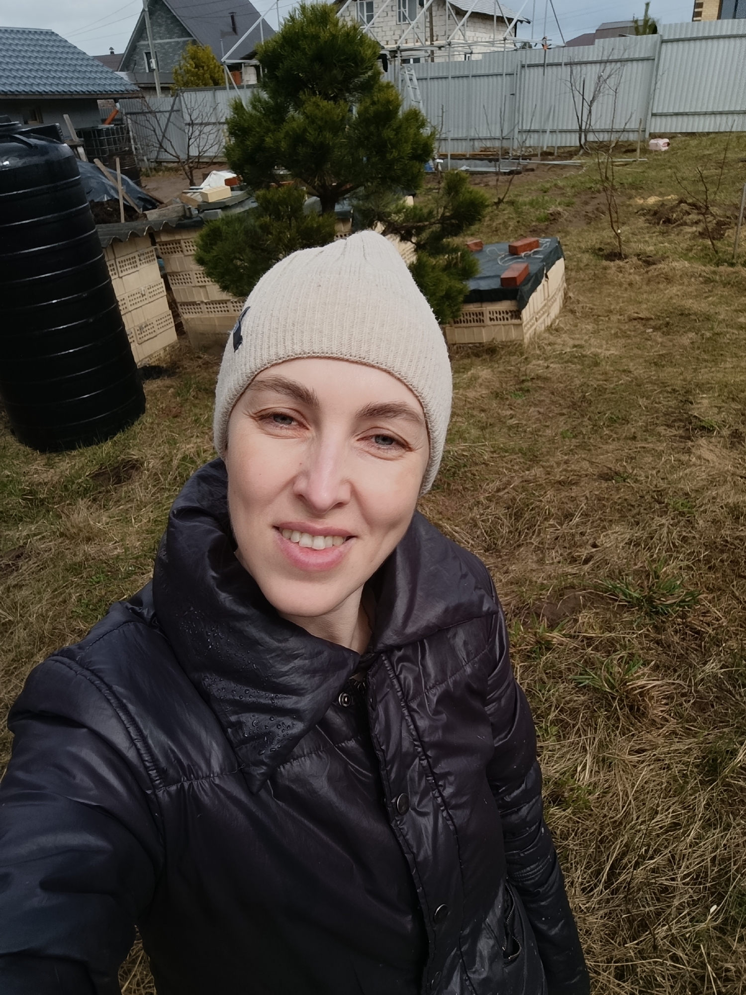 A smiling woman is captured in a selfie, set in what appears to be a residential backyard or garden in Chaikovsky, Russia. She is shown from the chest up, wearing a light beige knitted beanie and a dark, possibly black or navy, puffer jacket with visible water droplets, suggesting damp conditions or recent rain. Her eyes are blue, and she has a pleasant, happy expression.
The background reveals a somewhat unkempt outdoor space with dry, sparse grass and patches of bare earth. To the left, a large black plastic barrel or tank is prominent. Further back, light-colored bricks or hollow blocks are stacked, forming low structures. A small, bushy evergreen tree stands in the mid-ground, flanked by several bare deciduous trees or shrubs, hinting at a cooler season like early spring or late autumn. In the far background, a tall, corrugated metal fence stretches across, behind which parts of houses with grey roofs and light-colored walls are visible. The sky is overcast, providing diffused light and indicating a cloudy day. There is no discernible legible text within the image.