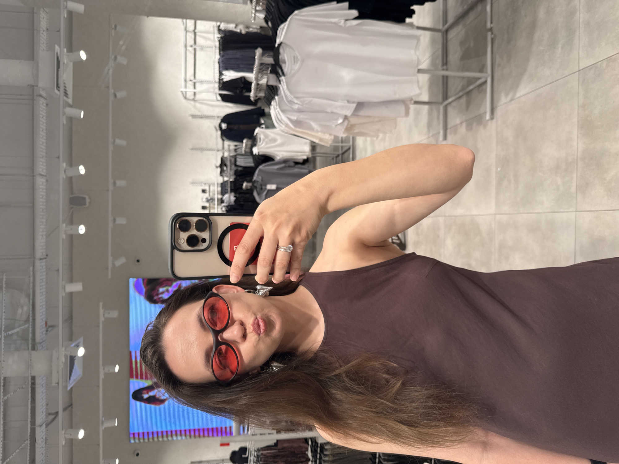 A woman takes a selfie in a clothing store, evident from the clothing racks behind her filled with garments. She is wearing red-tinted sunglasses and a brown tank top. Her hair is long and brown. She holds a smartphone in her right hand, with a red pop socket visible on the back of the phone. Her left hand is partially visible, showing a ring on her ring finger. The store appears to be well-lit with a modern interior. A large screen with colorful content is partially visible in the background. The woman is pouting her lips. The exact location in Khimki, Russia is not identifiable from the image.