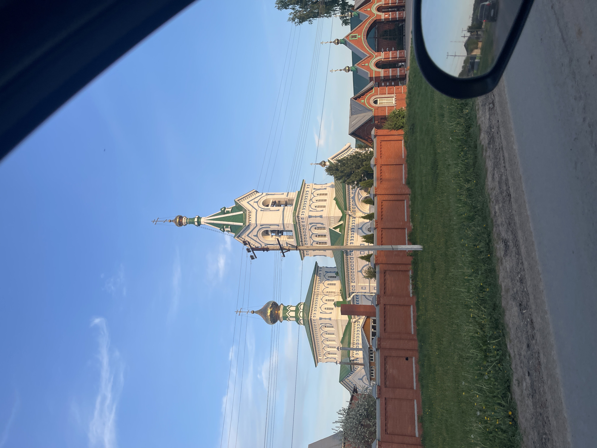 The image shows a view from inside a car, looking at a church complex in Перевозное, Russia. The complex includes two prominent white and green Orthodox churches with golden domes and a tall bell tower. A red brick fence runs along the perimeter of the property, separating it from a strip of green grass and the road. In the car's side mirror, additional buildings and power lines can be seen reflected. The sky is blue with some wispy clouds.
