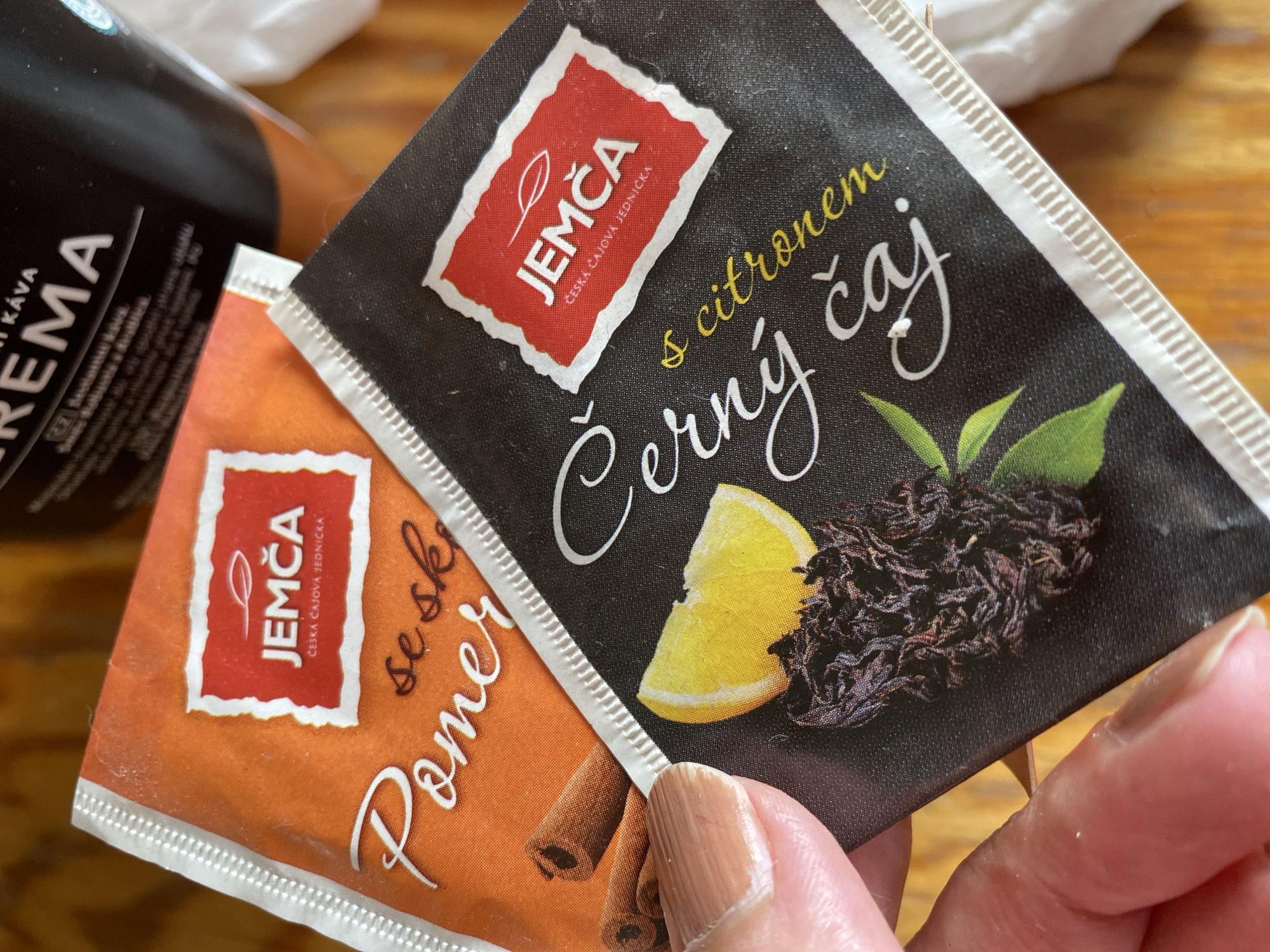 This is a close-up, overhead shot of two tea bags and part of a coffee container. The tea bags are the primary focus. The tea bag in the foreground on the right is black and features a red logo with "JEMČA" in white letters, along with smaller text below. Larger, white cursive text reads "Černý čaj" (black tea), and yellow cursive text above it says "s citronem" (with lemon). A slice of lemon and a pile of dark tea leaves are depicted on the black background.
To the left and slightly behind is an orange tea bag. It also has a red "JEMČA" logo with smaller Czech text. White cursive text on the orange bag reads "se sk Pomer" (likely part of a flavor name).
The coffee container to the far left is black with white lettering that appears to say "KÁVA CREMA".
A person's hand and fingernails are visible in the bottom right corner, holding the black tea bag. The background appears to be a wooden surface. The lighting suggests it could be daytime, possibly indoors. The overall impression is a casual moment, perhaps someone preparing tea or coffee. The text on the packaging is in Czech, indicating a likely location in the Czech Republic.