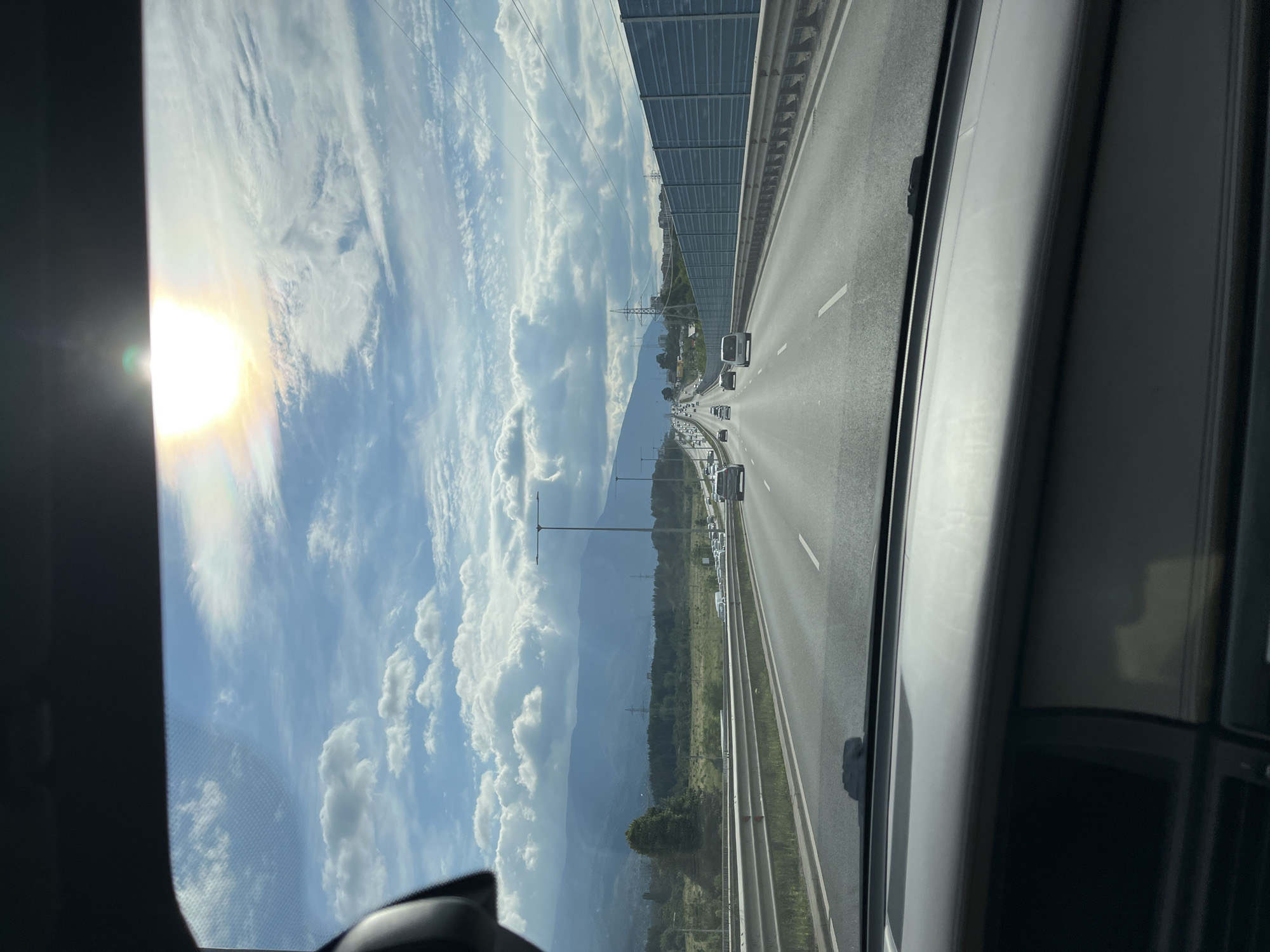 The image captures a view from inside a vehicle traveling on a multi-lane highway. The sun is visible high in the sky, partially obscured by clouds, casting a bright light. The highway stretches into the distance, with other vehicles on the road. To the left, there are mountains and trees, with power lines visible in the background. To the right, a sound barrier lines the highway. The dashboard and windshield of the vehicle are visible in the foreground. The location is identified as the "North-South Highway" in Sofia, Bulgaria.