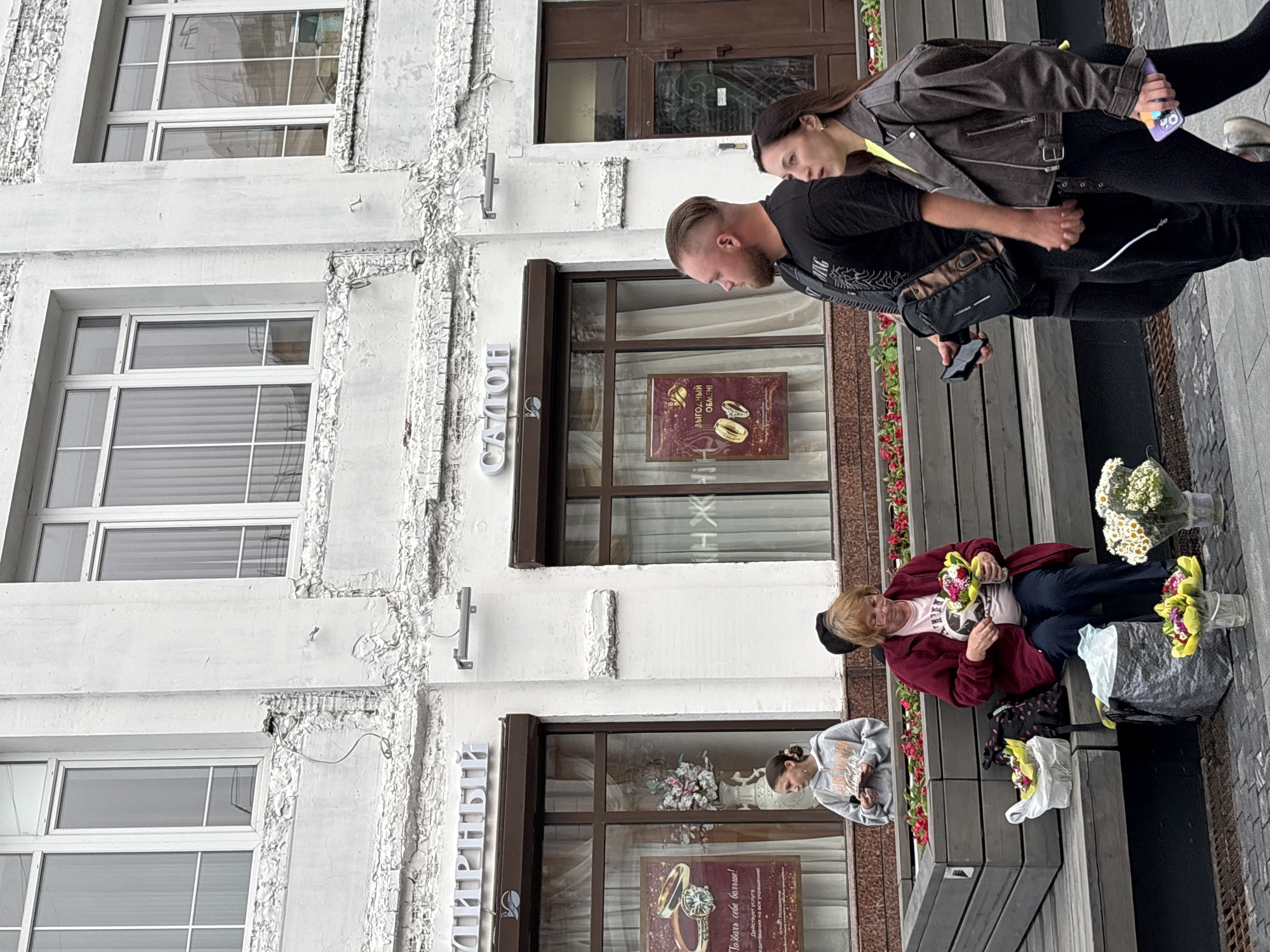 On a cloudy day in Nizhny Novgorod, Russia, in front of a white building with a textured facade and large windows, a street scene unfolds. The building houses establishments with signs reading "ТИРНЫЙ" and "САЛОН".
In the foreground, a dark grey wooden planter box runs along the front of the building, filled with red flowers. Seated on a bench in front of the planter is an older woman wearing a dark red jacket and a light pink t-shirt. She is holding a bouquet of flowers and appears to be arranging them. To her left, on the bench, are two bags, one black with a polka dot pattern and another white plastic bag, both containing flowers. On the ground in front of the bench, several more bouquets of flowers are arranged in small buckets, some wrapped in yellow paper.
To the right of the seated woman, a young woman in a grey hooded sweatshirt sits on the bench, holding a small item in her hands.
Further to the right, a couple walks past. The man, with a shaved head and a beard, wears a black t-shirt and a dark messenger bag slung across his chest. He is holding a black smartphone in his left hand. The woman beside him wears a brown leather jacket over a bright yellow top and black leggings. She is holding a purple smartphone in her right hand and appears to be walking while looking ahead.
The ground in front of the planter and bench is paved with grey slabs, with some visible drain grates. The overall atmosphere suggests a commercial street with flower vendors.