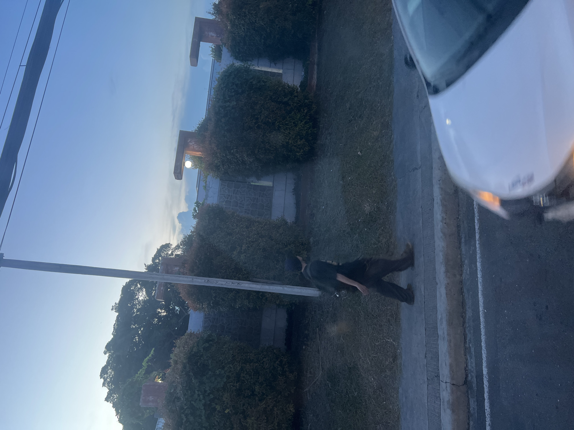 A person walks along a sidewalk in Davao City, Philippines, with a white car in the foreground. The person is wearing a dark t-shirt and brown pants. They are walking from left to right across the image. Behind them are manicured bushes and a stone wall, with a street light visible above the bushes. The sky is a light blue, with some clouds. The image appears to have been taken at dusk.