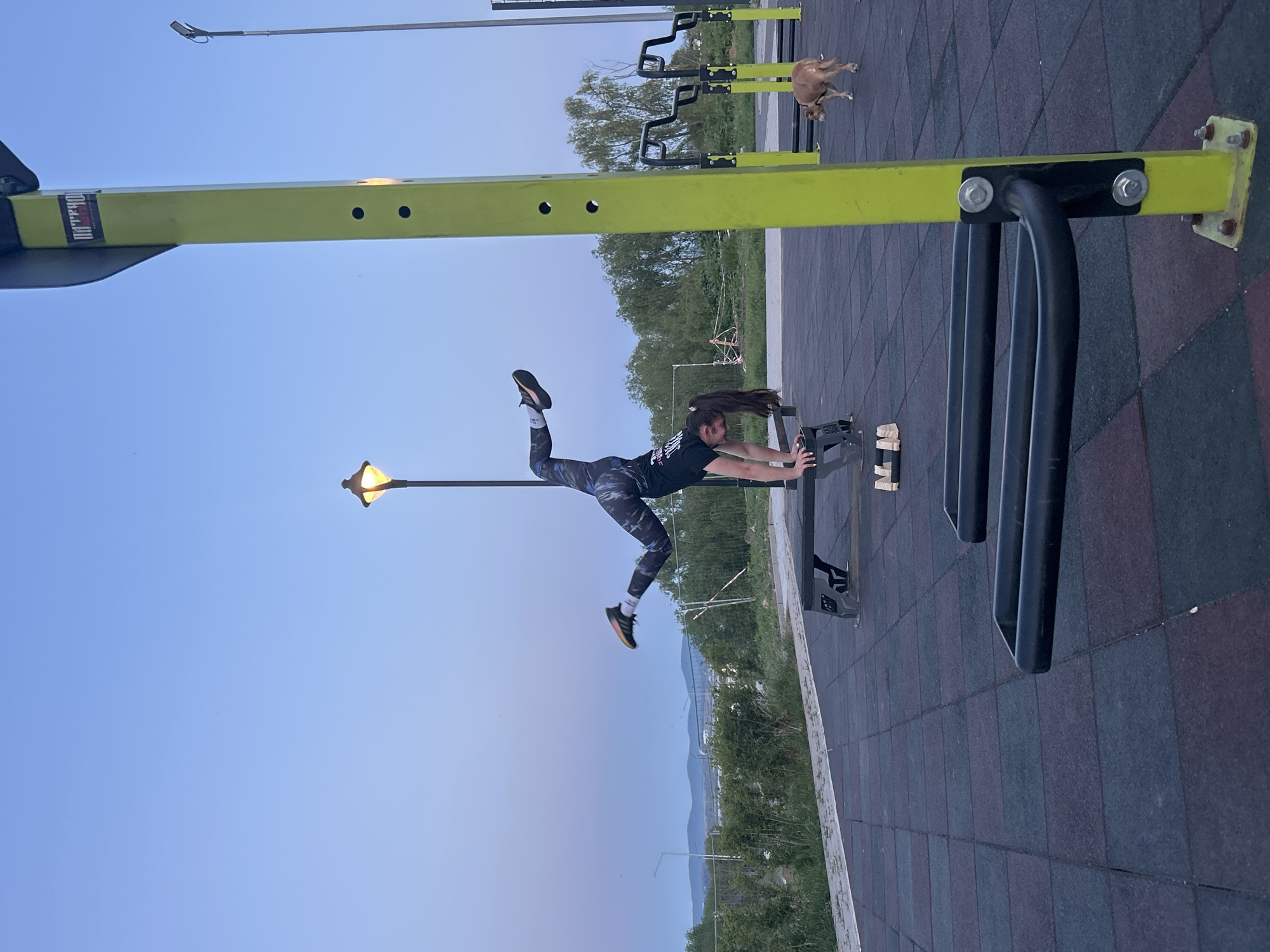 The image captures a woman performing a dynamic stretching exercise on an outdoor exercise bench in a park-like setting. She is wearing a black t-shirt, patterned leggings, and athletic shoes, and is positioned horizontally, gripping the sides of the bench while extending her legs upwards. A street lamp stands to her left. In the background, other exercise equipment is visible. A dog is present in the scene. The scene is located in the city of Burgas, Bulgaria. The ground is covered with a rubberized tile surface. Trees and distant mountains are visible in the background. The sky is a light blue, suggesting either early morning or late evening.