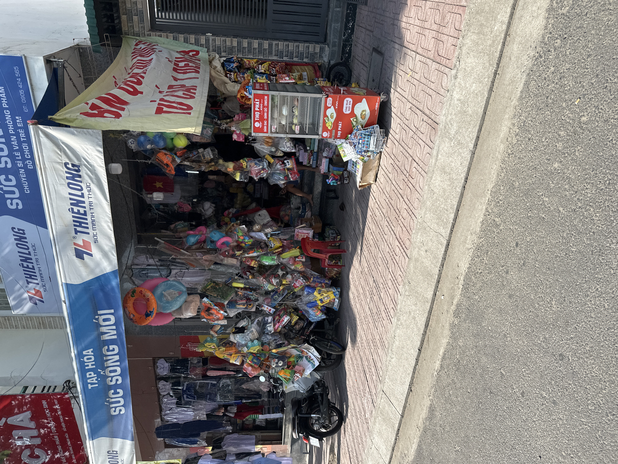 The image depicts a street scene in the city of Bac Nha Trang Ward, Vietnam, featuring a storefront that appears to be a small general store or convenience shop. The shop displays a wide assortment of goods, including clothing, toys, and possibly food items, which are densely hung and arranged both inside and outside the establishment. A motorcycle is parked in front of the shop, adding to the cluttered but vibrant atmosphere. A red plastic chair is placed on the sidewalk near the entrance. A display case with food items, possibly steamed buns, is positioned on the sidewalk. The storefront has prominent signage, including a blue banner with Vietnamese text. The street is paved with asphalt, and the sidewalk is made of patterned tiles. The overall impression is one of a busy, local commercial area.