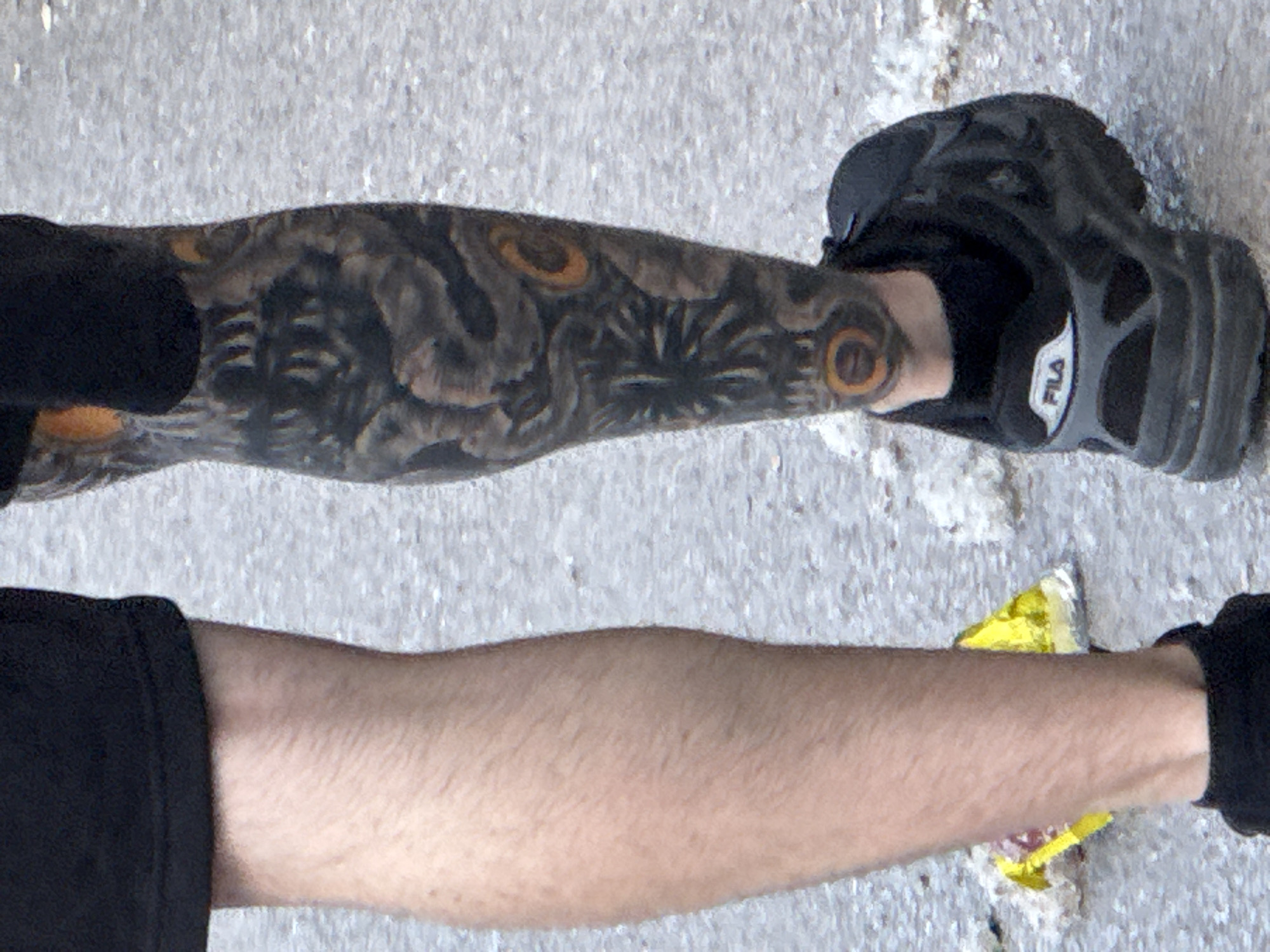A photograph shows two legs on a light grey surface. The upper leg has a dark tattoo with circular, eye-like designs. The foot on that leg is wearing a black Fila sneaker. The lower leg is bare. The location is unknown and cannot be determined from the image. No interactions or actions are visible.