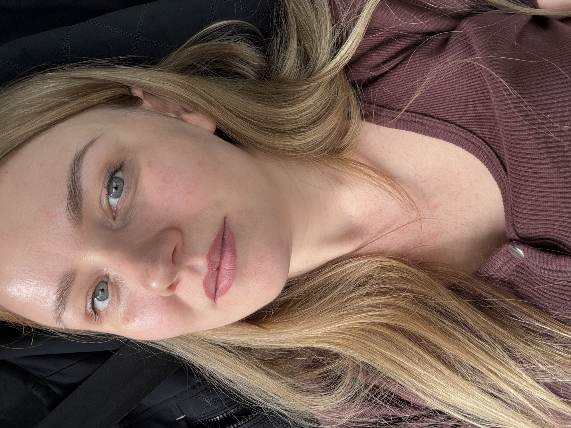 A portrait-style close-up photo shows a woman with long, light blonde hair and blue-grey eyes, looking directly towards the camera. She appears to be lying down, with her head tilted to her right. Her hair is spread around her shoulders and against a dark, quilted surface, possibly a jacket or seat.
The woman has a calm, neutral expression. Her eyebrows are neatly shaped, and she wears subtle cheek blush. Her lips are a soft mauve-pink, exhibiting a crisp and defined outline that suggests freshly applied lip blush or permanent makeup. She is wearing a maroon-colored, ribbed top or sweater. The lighting is soft, hinting at an indoor setting or diffused daylight. No specific time of day or weather can be discerned. There is no visible text in the image. This scene takes place in Izhevsk, Russia.