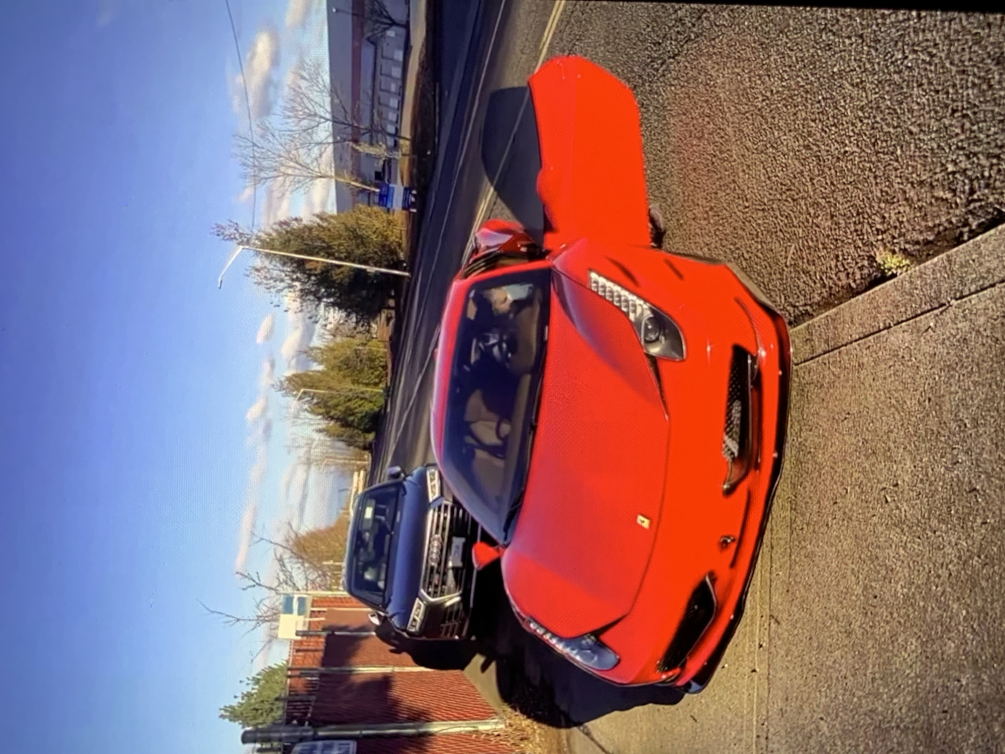 A bright red Ferrari is parked on a paved street next to a sidewalk. The driver's side door of the Ferrari is open. Behind the Ferrari, a dark-colored Audi SUV is parked on the street. The vehicles are in front of a chain-link fence. The sky is clear and blue with some scattered clouds. The sun is casting long shadows on the ground. Trees and buildings are visible in the background.
