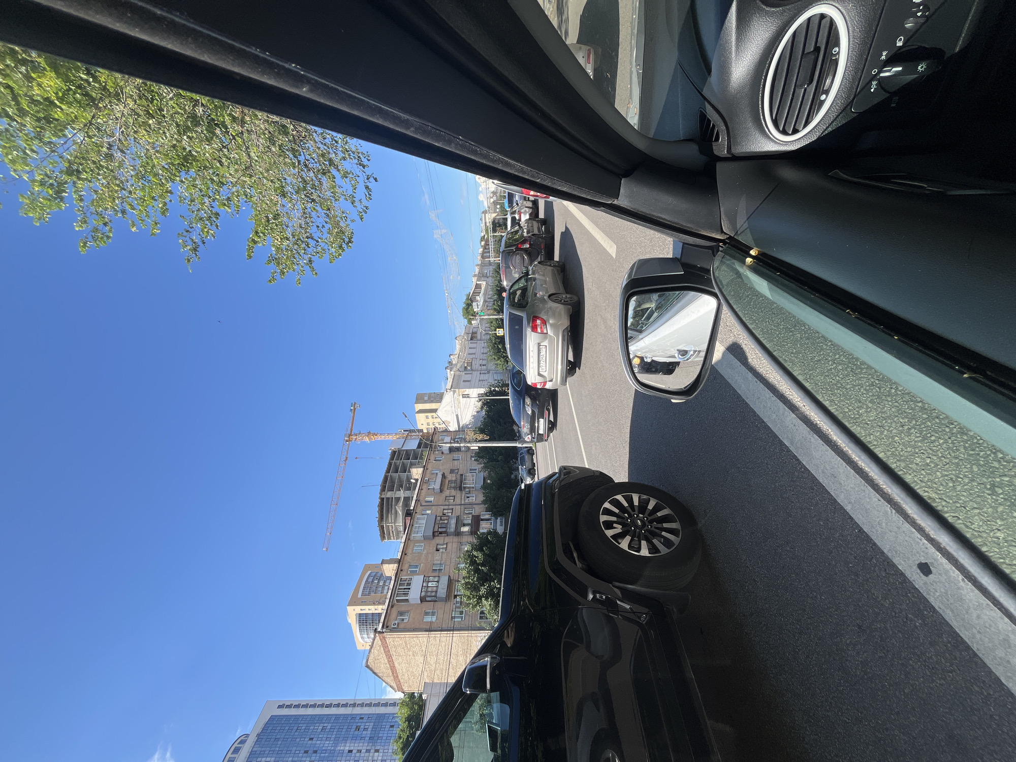 This media file captures a view from inside a car, looking out the driver's side window and through the windshield. The scene depicts a traffic jam on a sunny day. Several cars are visible in the lane ahead, indicating a slowdown or complete stop. The surrounding environment suggests an urban setting with buildings lining the street. A construction site with a crane is noticeable in the background, implying ongoing development in the area. The sky is clear and blue, suggesting fair weather. The sun is casting shadows, consistent with daytime. The perspective suggests the vehicle is either parked or moving slowly in traffic. No people are clearly visible within the frame. The image also shows parts of the car's interior, including a side mirror and dashboard elements like an air vent and controls.