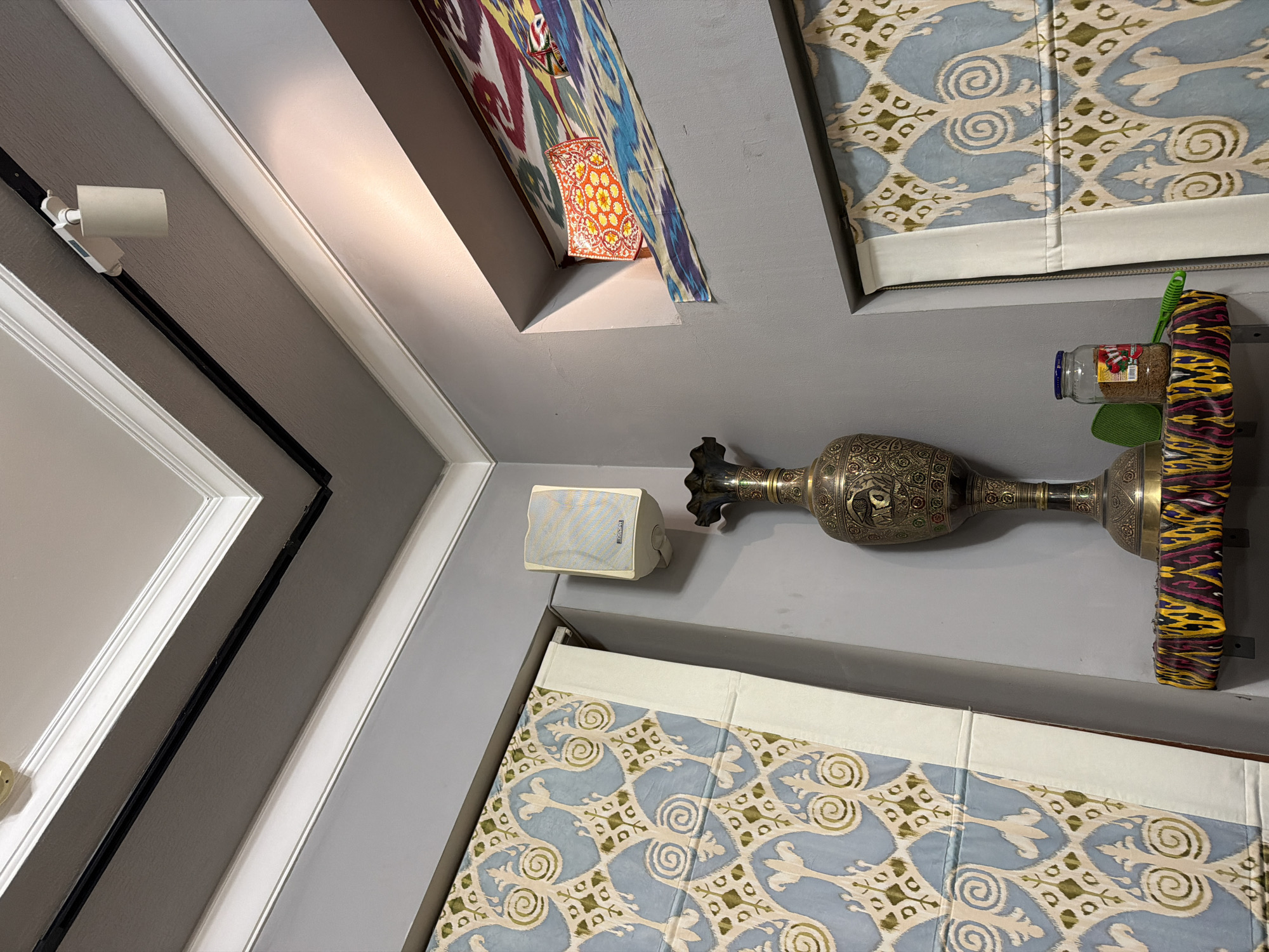 The image captures an indoor corner with gray walls. On the left, a patterned curtain with blue, beige, and green motifs hangs vertically. To the right of the curtain, a white speaker is mounted on the wall. A tall, ornate brass vase sits on a shelf covered with a colorful, patterned fabric. Next to the vase is a glass jar containing a granular substance, possibly seeds or spices, with a green brush resting beside it. Above the vase, a recessed shelf displays a red decorative plate with yellow floral patterns and a colorful textile with abstract designs. A track light is visible on the ceiling, casting light downwards. The overall impression is of an interior space decorated with traditional Uzbek textiles and decorative items.