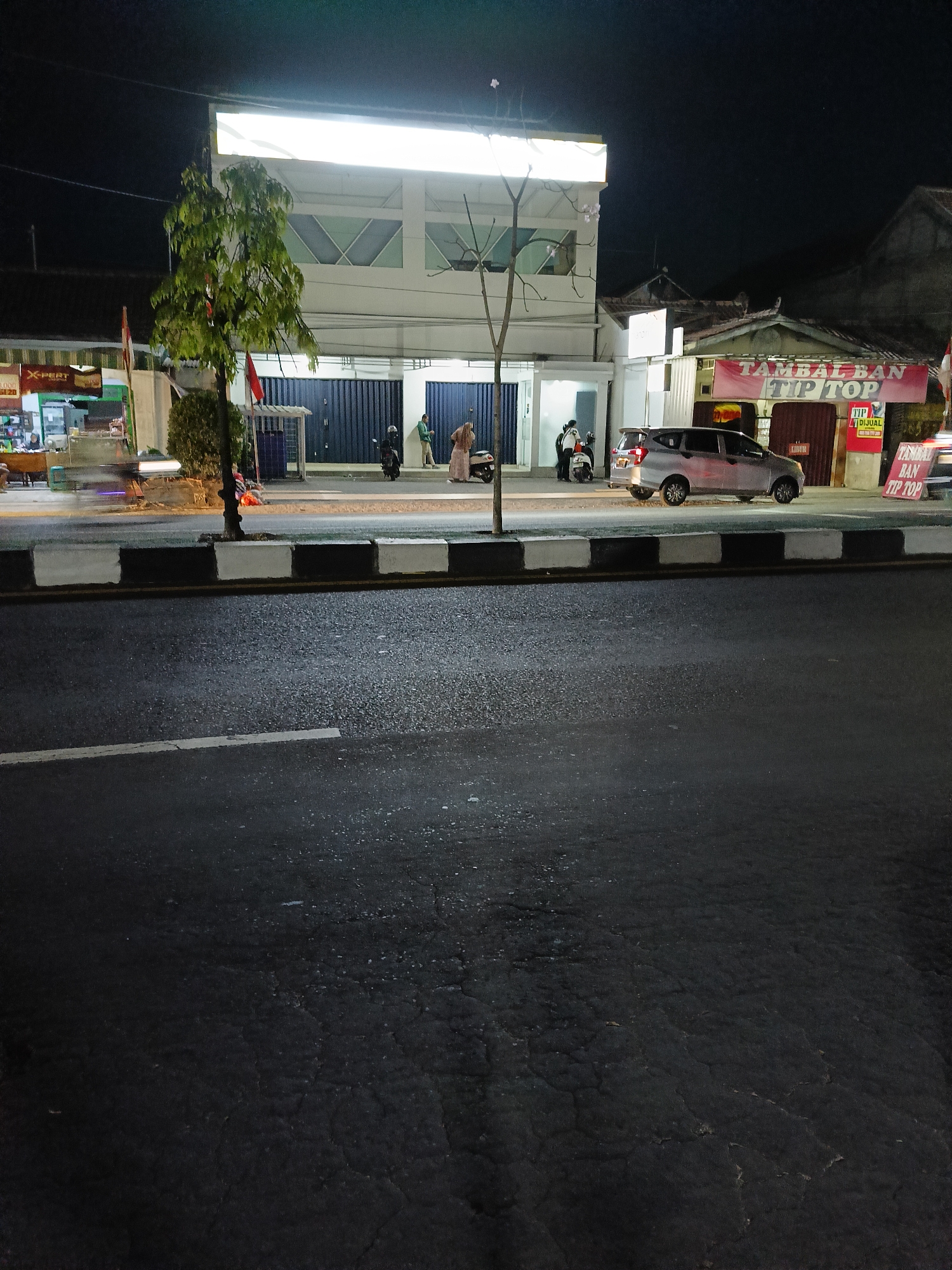 The image shows a street scene at night in Kebondalem Kidul, Indonesia. The foreground is dominated by a dark asphalt road with white and black striped curb markings. In the middle ground, a silver hatchback car is parked on the side of the road in front of a building with a banner that reads "TAMBAL BAN TIP TOP". There are several motorcycles and two individuals, one wearing a green shirt and the other a headscarf, standing near the motorcycles and the building. A tree with green leaves stands to the left of the building, and a blurry figure on a motorcycle is visible moving along the road in the far left of the image. The background consists of buildings and the night sky.