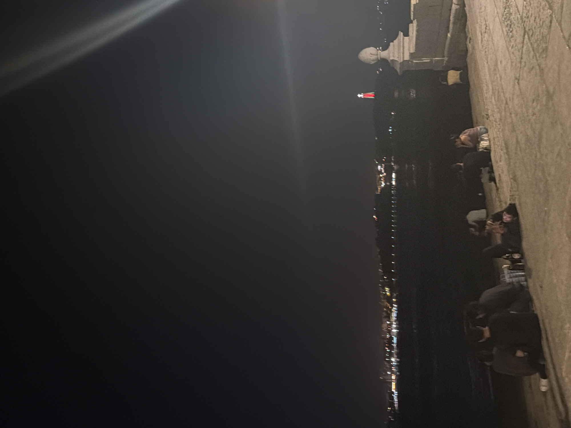This image depicts a night scene from the esplanade of Cais das Colunas in Lisbon, Portugal. Several individuals are seated on the ground, appearing to socialize and relax. In the foreground, a group of people are gathered on a paved area, with some bottles of drinks visible. Further back, other individuals are also seated, some appearing to interact. Across the Tagus River, the city skyline is illuminated by distant lights, and a prominent monument, likely the Cristo Rei statue, is visible in the distance, lit with red and green colors. The sky is dark, with some light beams suggesting artificial lighting in the upper right quadrant of the image. The overall atmosphere is one of a casual evening gathering by the waterfront.